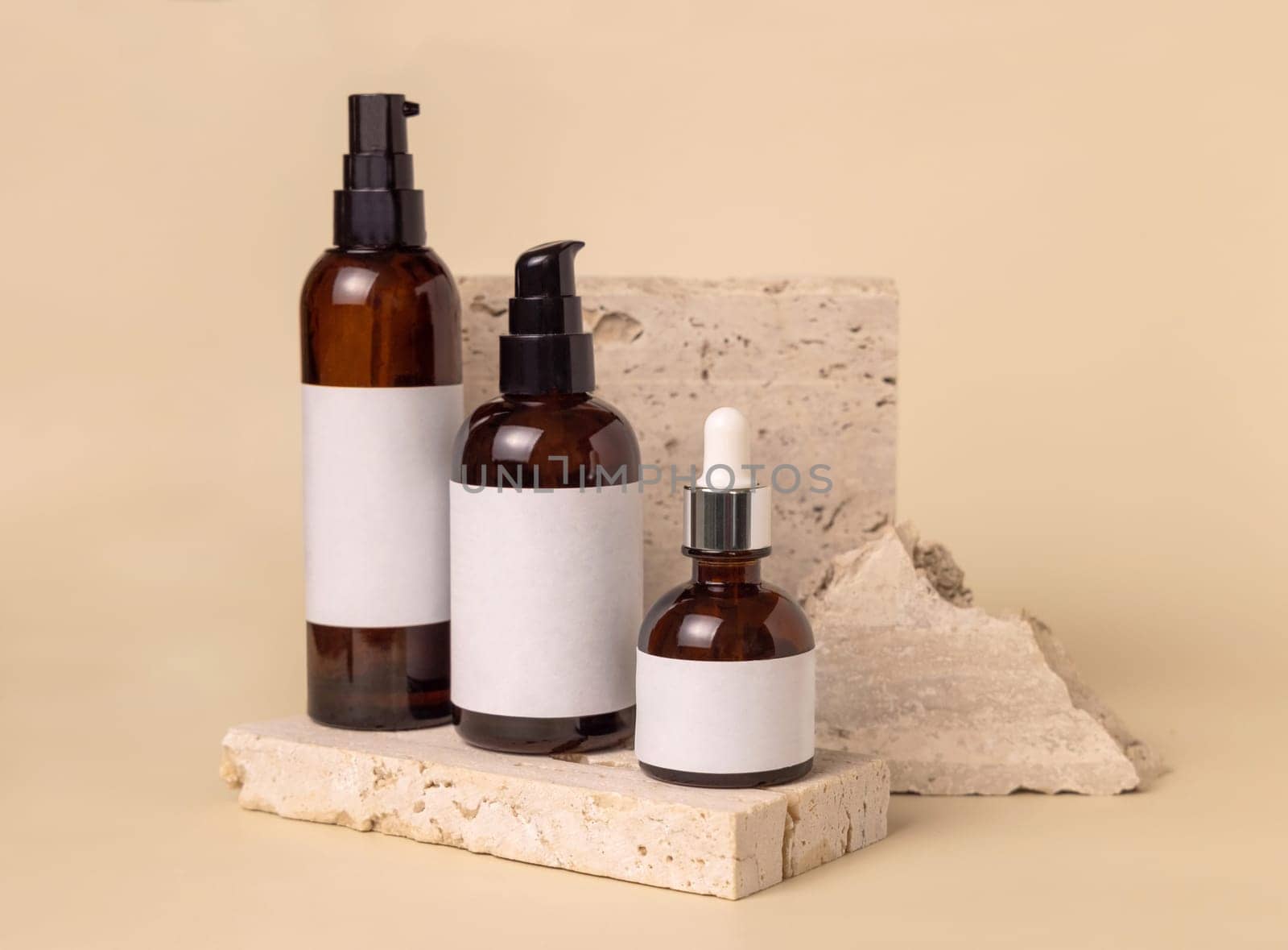 Brown glass one pump bottles on travertine stones on light beige close up, label mockup. Skincare beauty product package. Natural cosmetics, minimal composition..