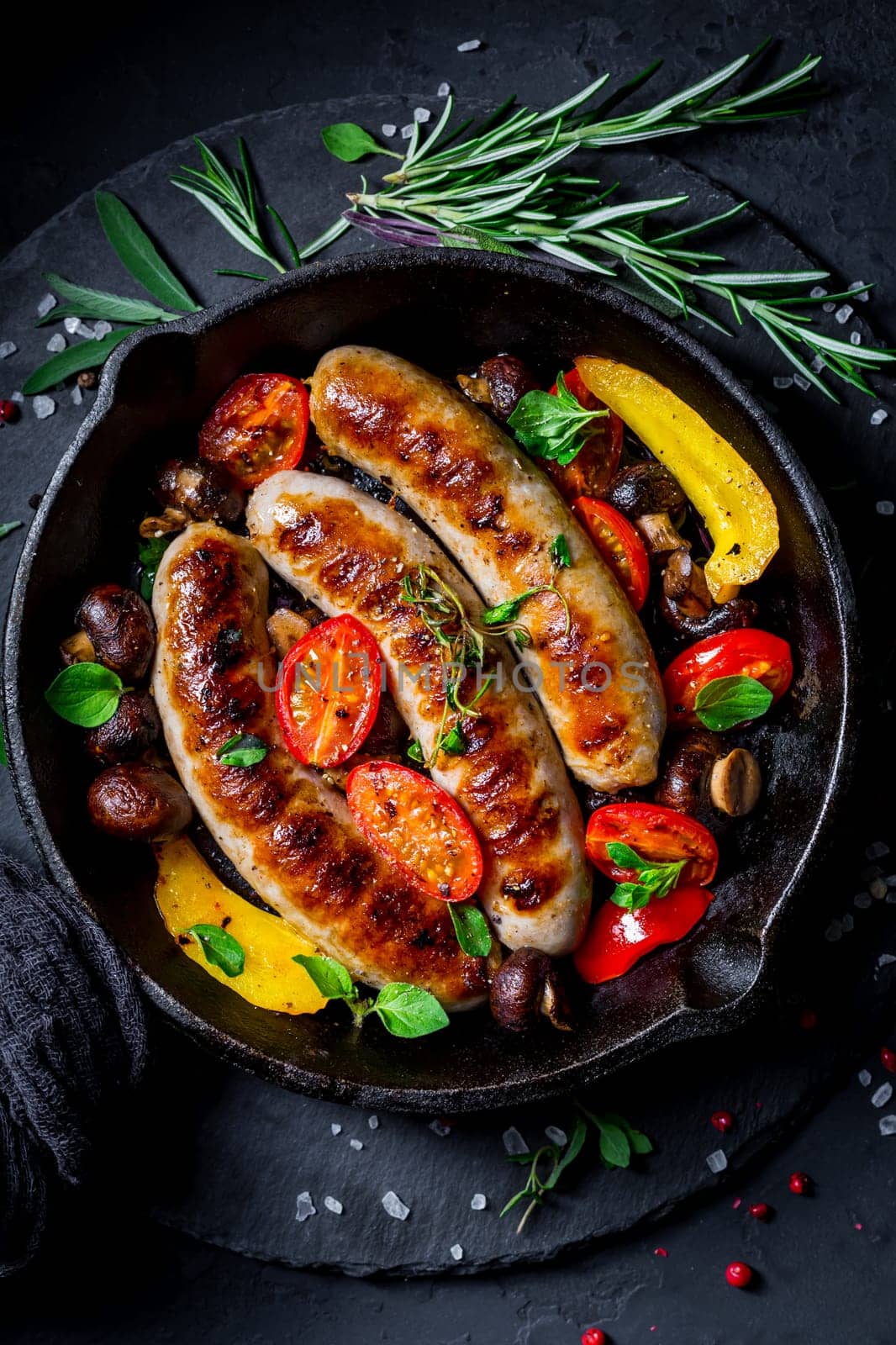 Grilled sausages and vegetables with herbs in iron pan on black background