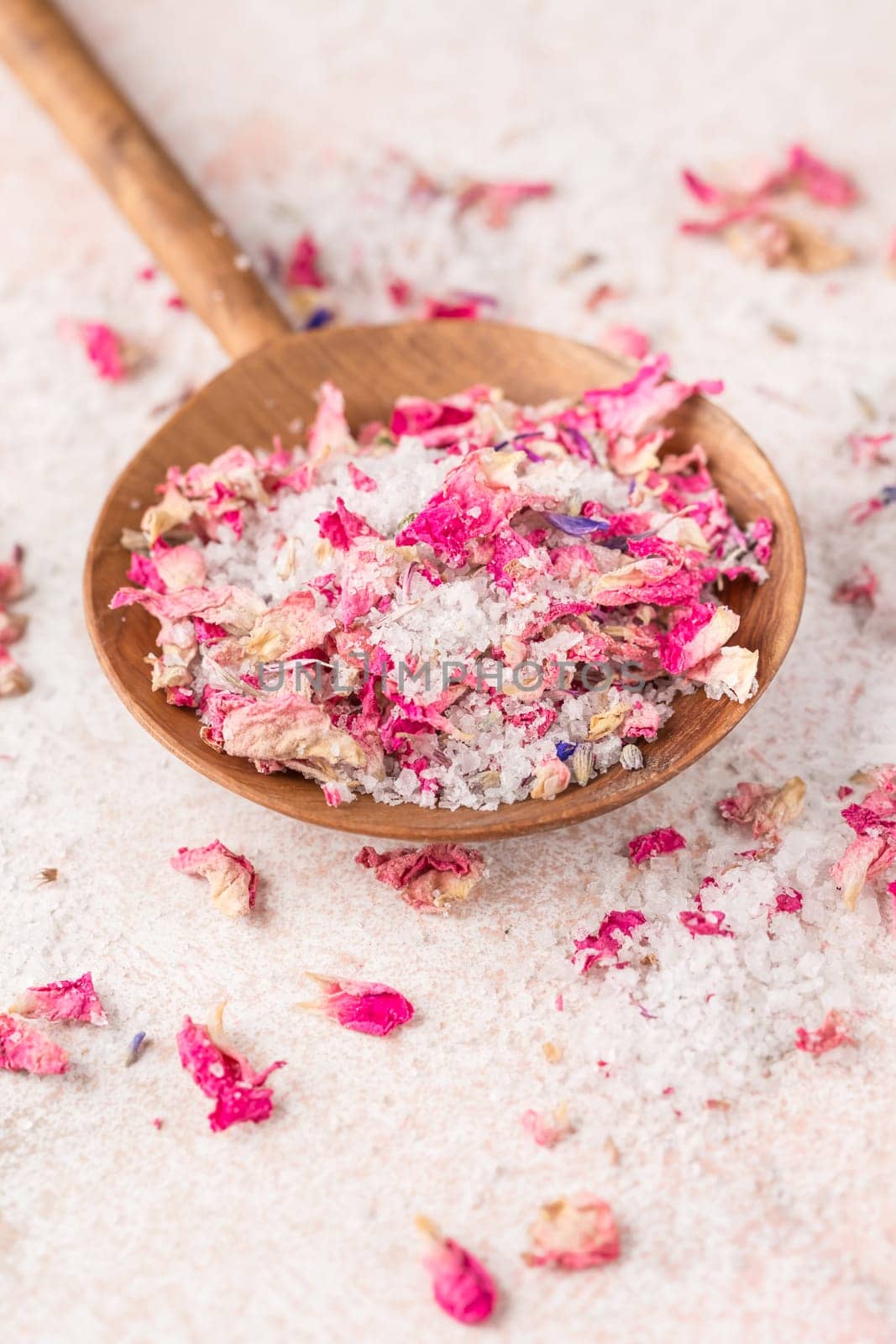 See salt wih with dried flower petals in wooden scoop on pink marble table