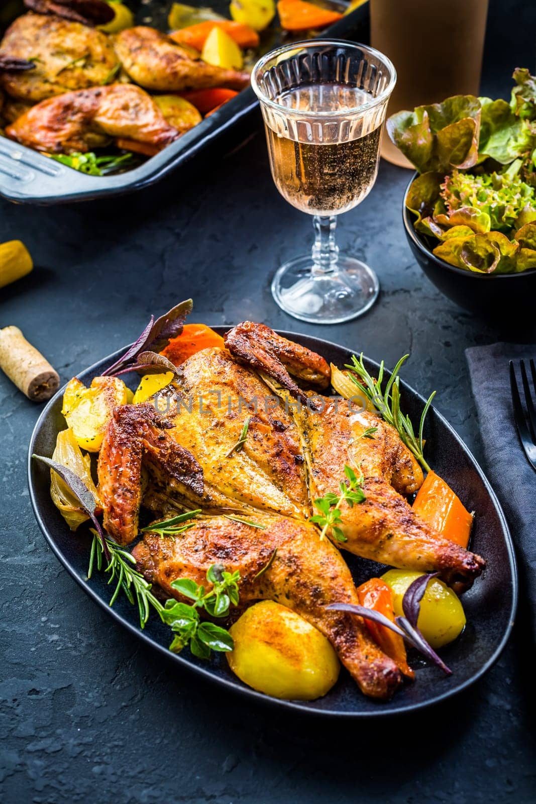 Baked whole chicken with potatoes, carrots, herbs and salt and rose wine on black background