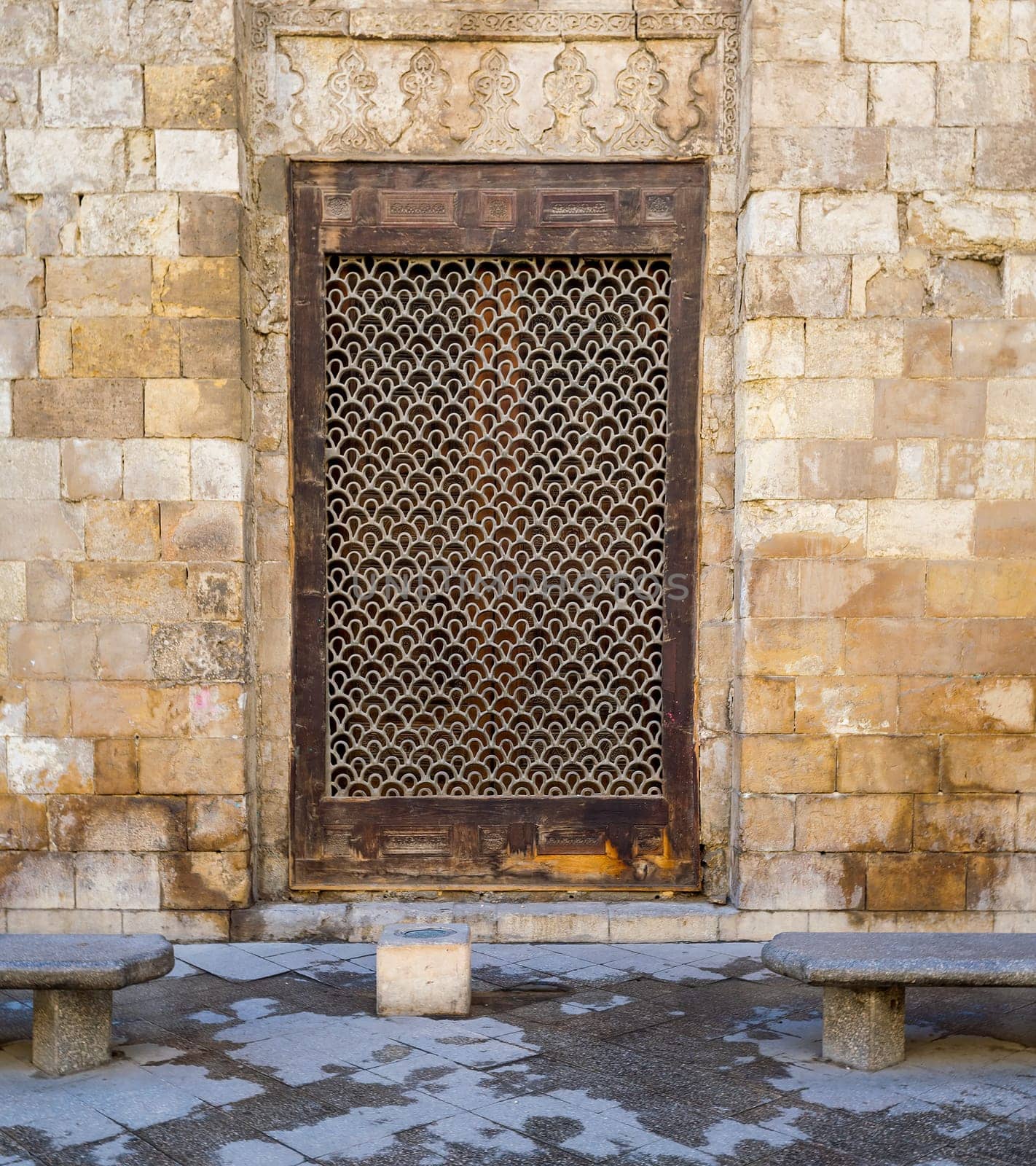Wooden grunge window with decorated iron grid over stone bricks wall and two marble garden benches, Moez Street, Cairo, Egypt