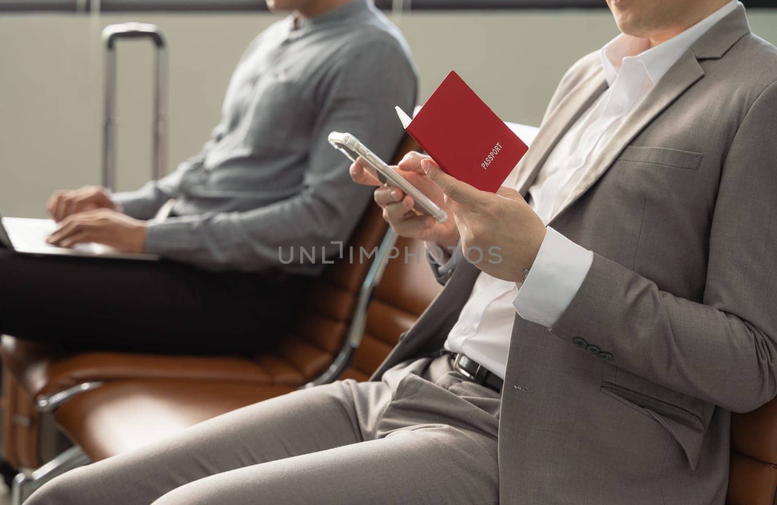 Businessmen waiting for flight in airport terminal. Man in suit holding passport sitting and working on mobile phone while other guy working on laptop. Business travel concept.