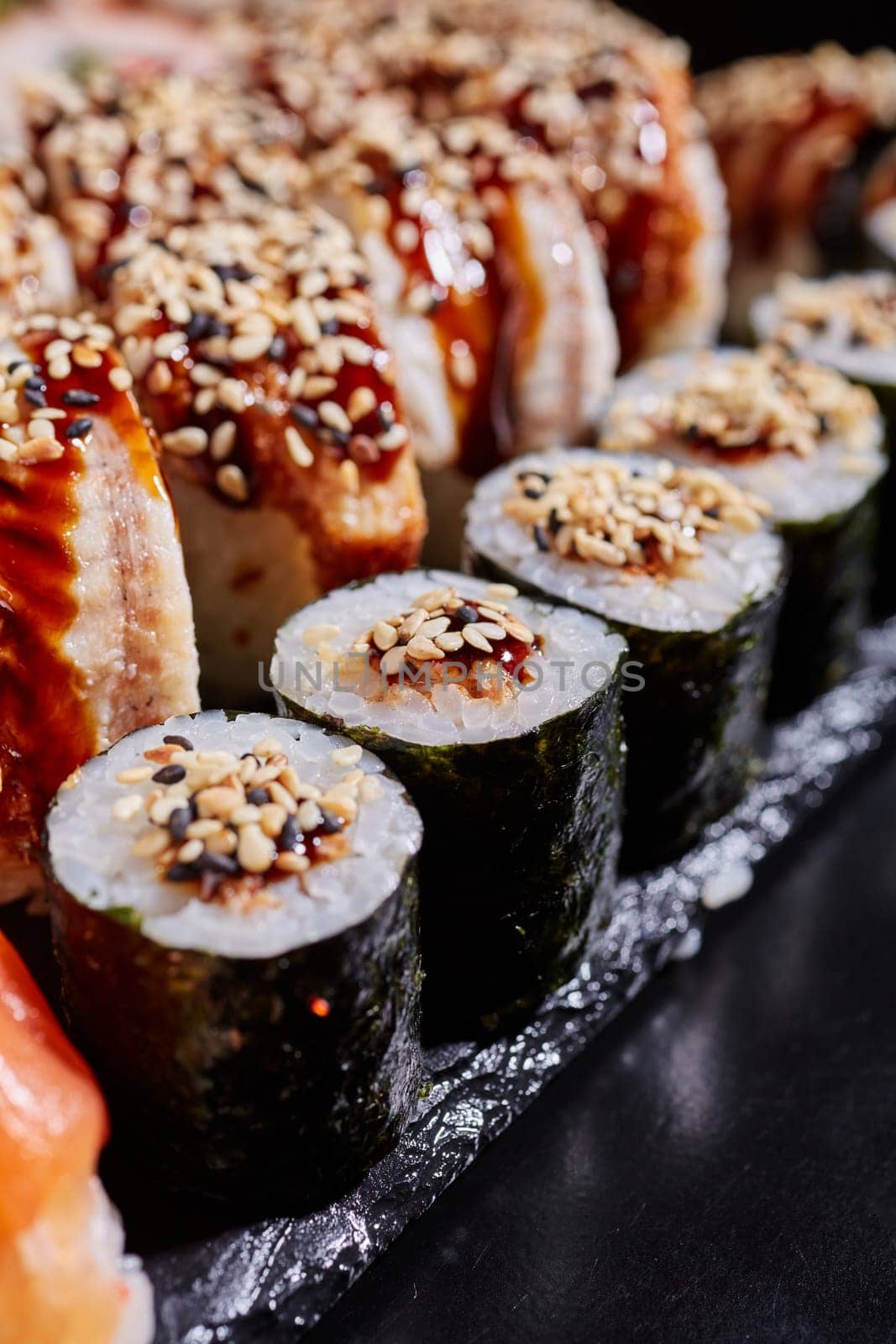 Close up of maki sushi with tuna, sauce and sesame seeds lying on the edge of a black stone plate. Selective focus. Blurred background.