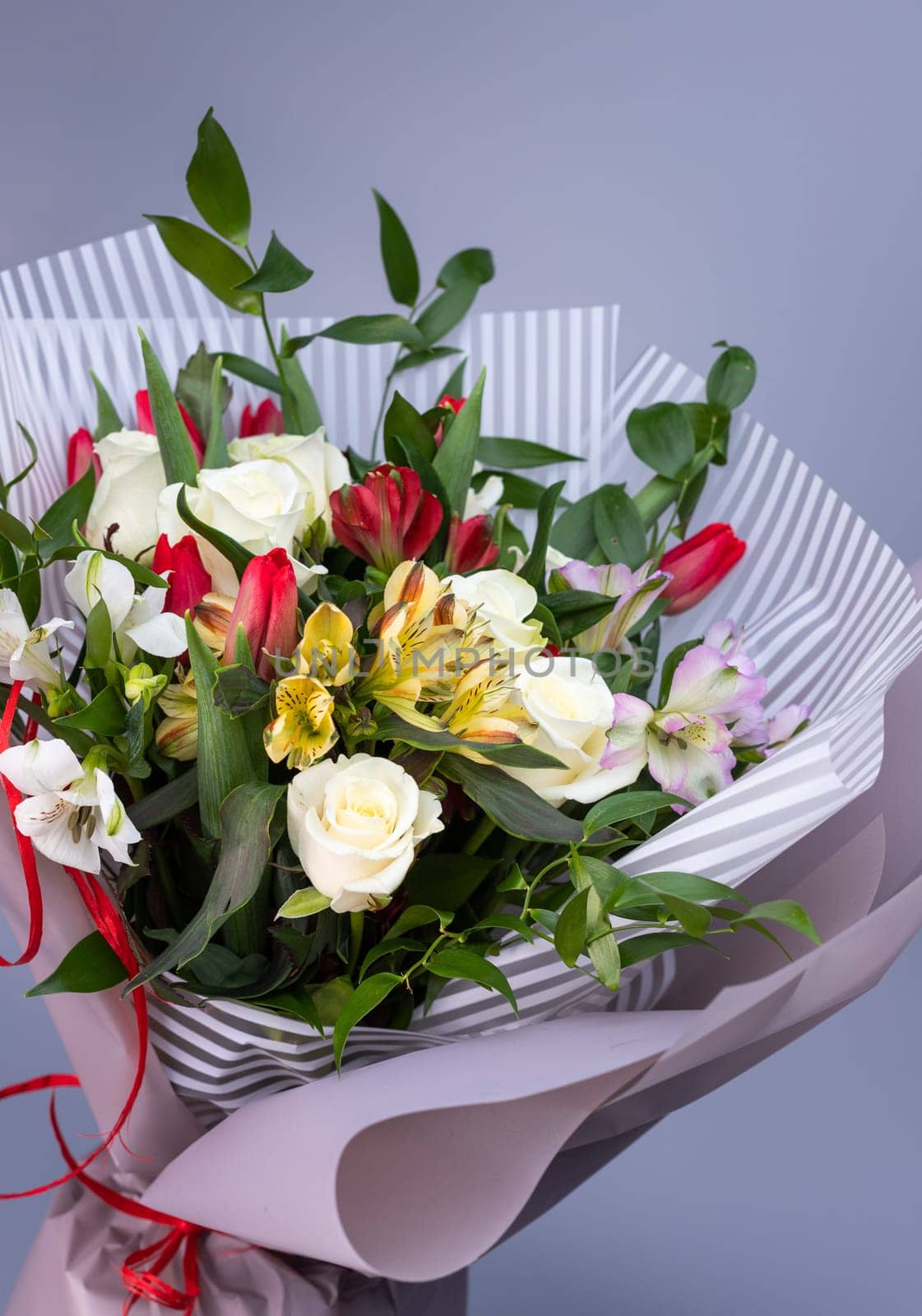 Close up of a bright bouquet of white roses, colored irises and red tulips. Flower bouquet wrapped in light floral paper of two kinds on a blue background. Background for greetings and postcards.