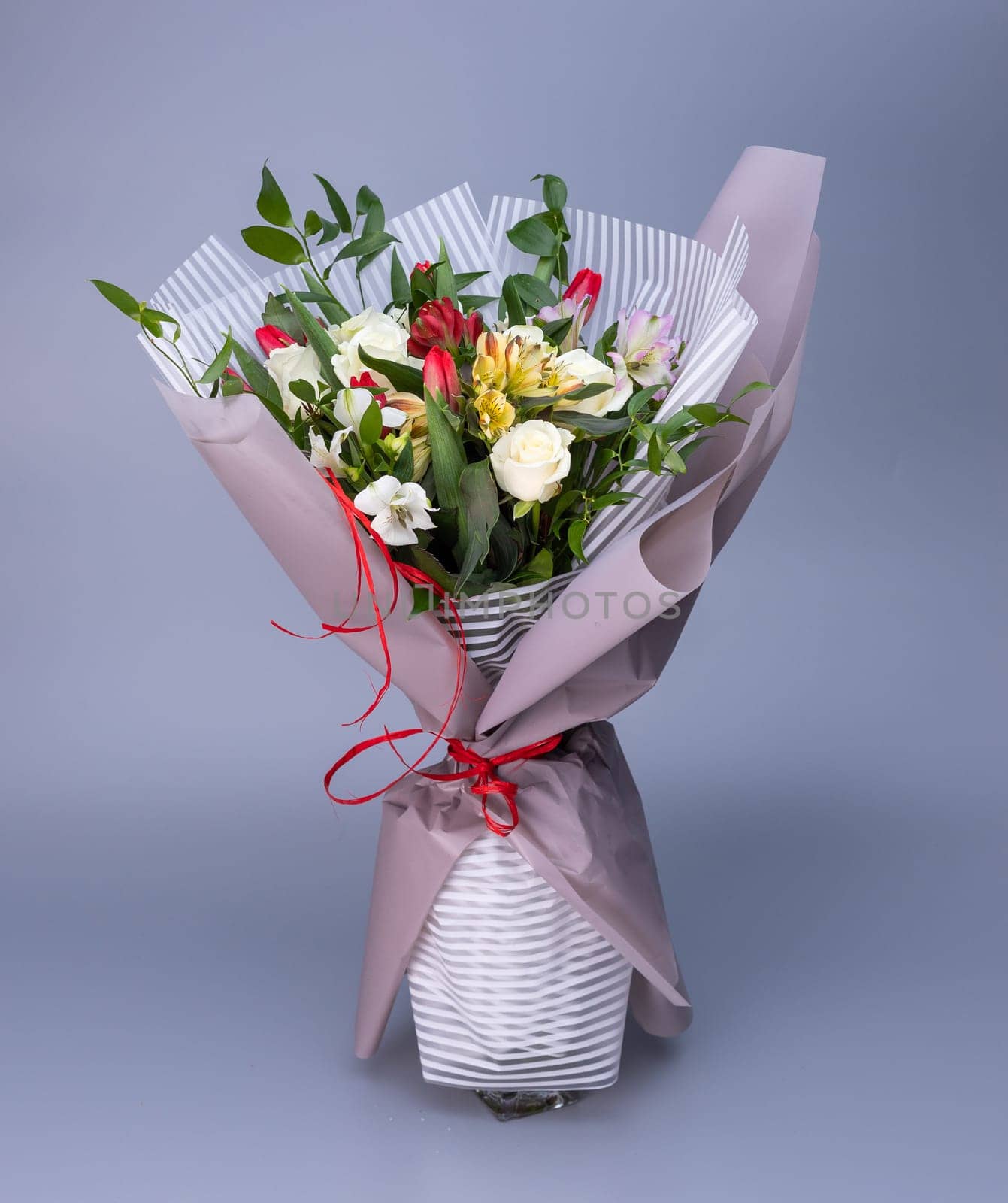 Flower bouquet, which is wrapped in light floral paper of two types stands on a blue background. Bouquet of white roses, colored irises and red tulips. Concept of floral arrangements for greetings.