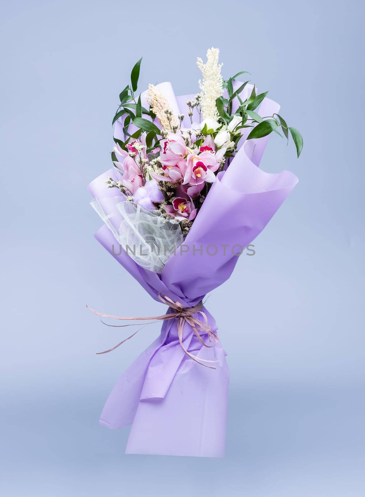 Bouquet of fresh natural flowers wrapped in floral tracing paper stands in a vase on a blue background. Bouquet in pink style composed of irises, other different flowers and twigs of cotton.