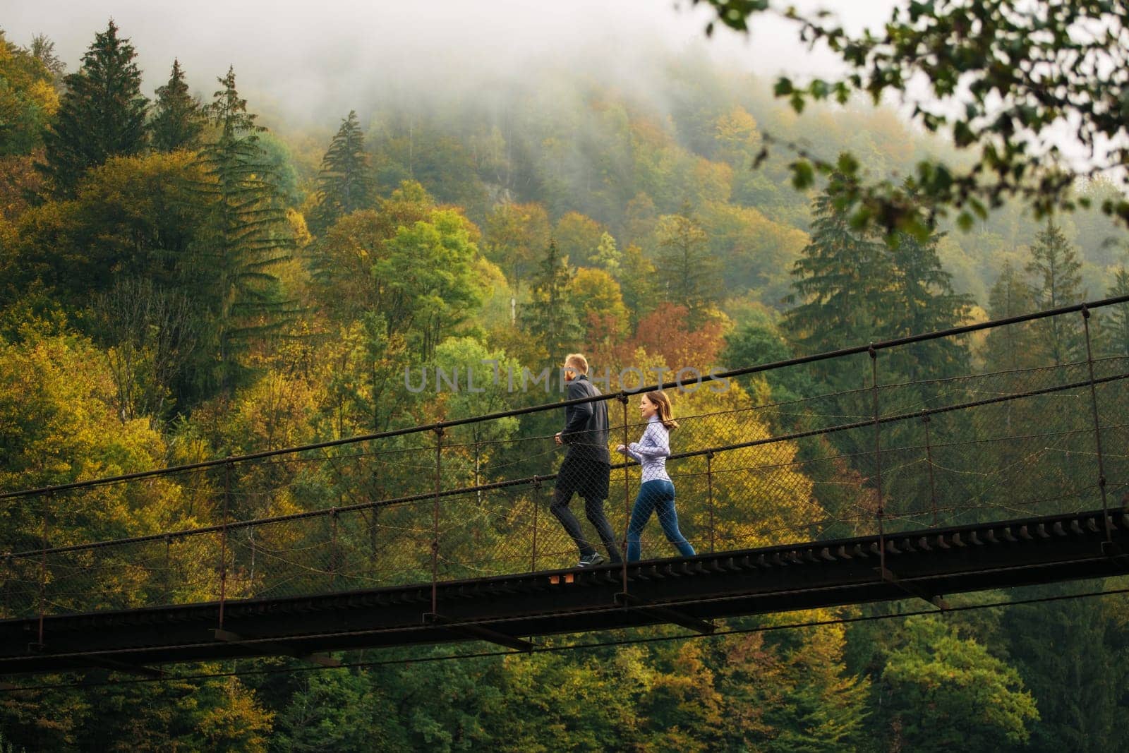 Living healthy lifestyle. View from below of a young athletic man and woman performing a morning run on a suspension bridge against the backdrop of a mountain forest. Mountain landscape.