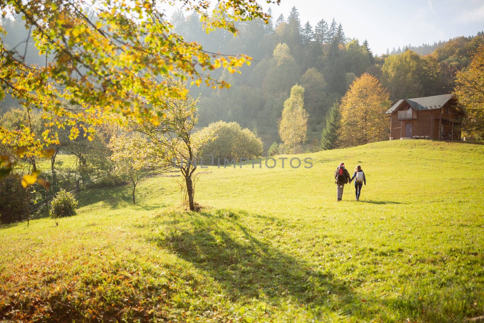 Two young tourists with backpacks on their backs holding hands head to a wooden house standing in the mountains. Concept of hiking, healthy and active lifestyle in young people.