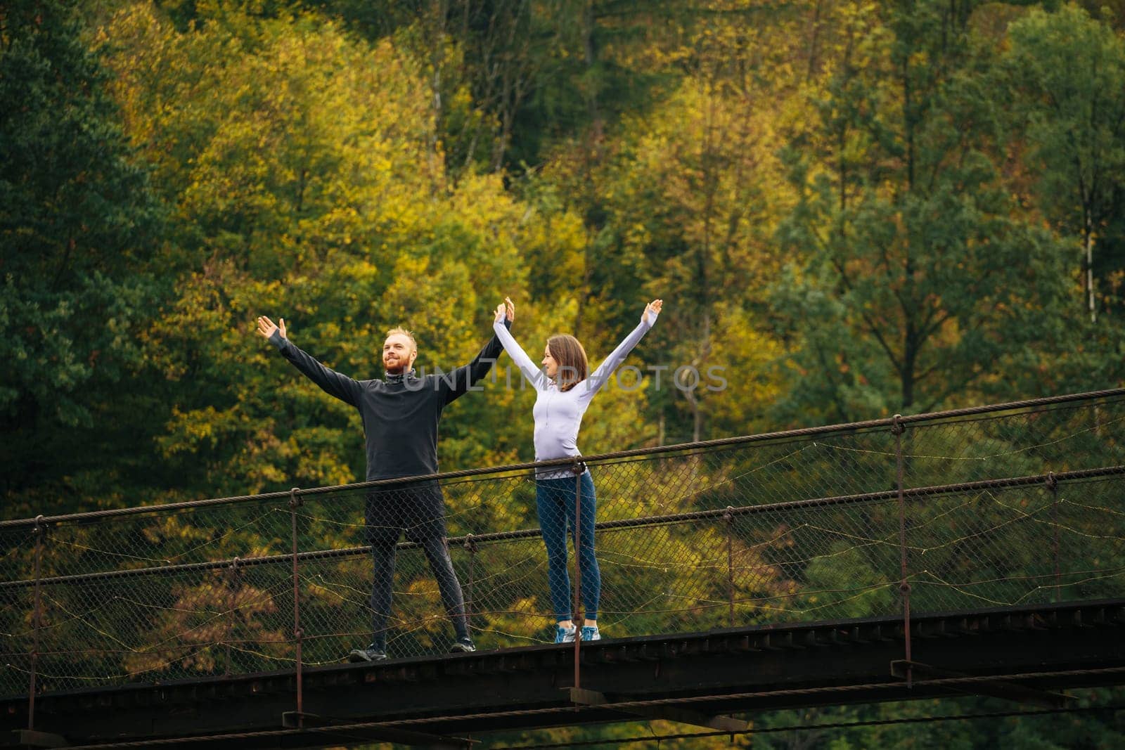 Healthy lifestyle. Young man and woman standing with arms raised on a suspension bridge on a background of forest. Sports couple warms up before jogging in the mountains.