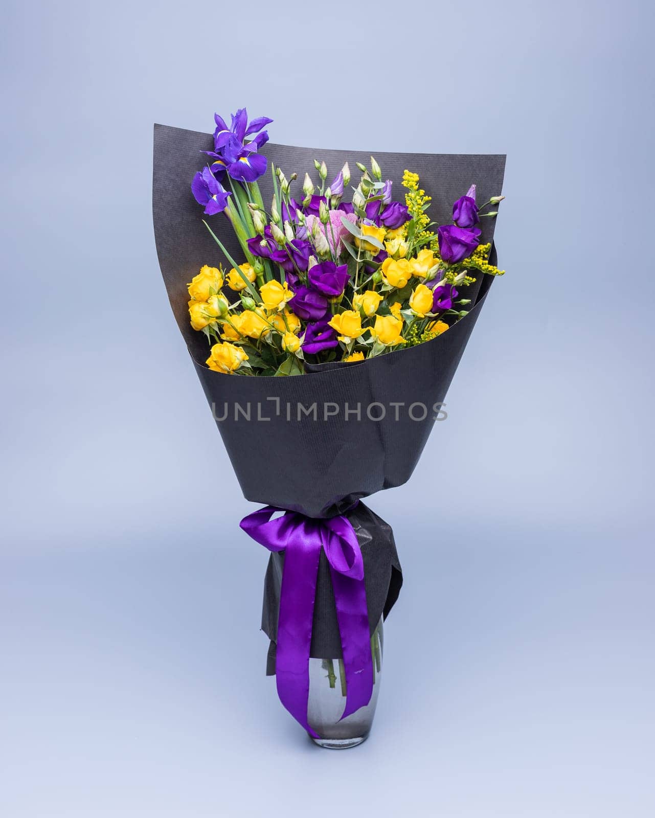 Stylish bouquet of yellow and purple flowers wrapped in black paper, stands in a vase on a blue background. Festively packed bright colored bouquet of yellow and purple eustoma and irises.