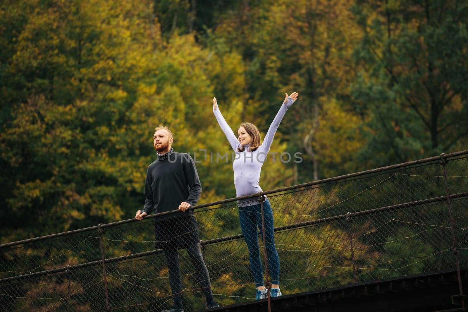 Mountain forest landscape. Young happy man and woman with joyful facial expression standing on a suspension bridge. Athletes in sports uniform are going to train outdoors. Concept of unity with nature