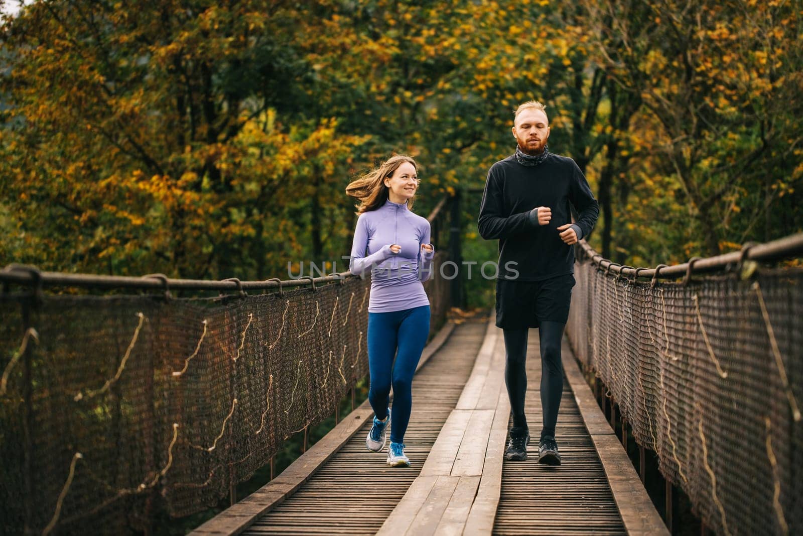 Young family spends time with benefit. Healthy sports couple in love running together on a wooden suspension bridge against the backdrop of the forest. Two runners on a morning jog.
