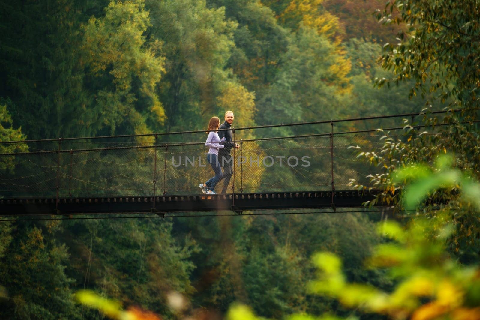Mountain landscape. Side view of a young man and woman jogging on a mountain suspension bridge. Athletes in sportswear train outdoors. Young family spends time with benefit.