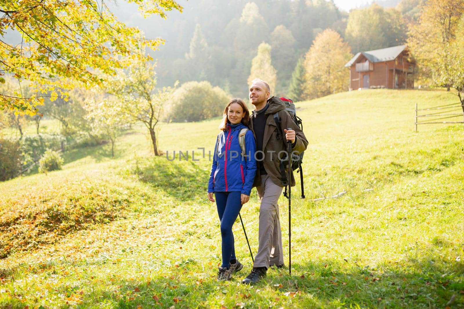 Portrait of a couple of young hikers with backpacks and trekking stick on a background of mountain countryside. Concept of hiking, tourism, nature, people and lifestyle.