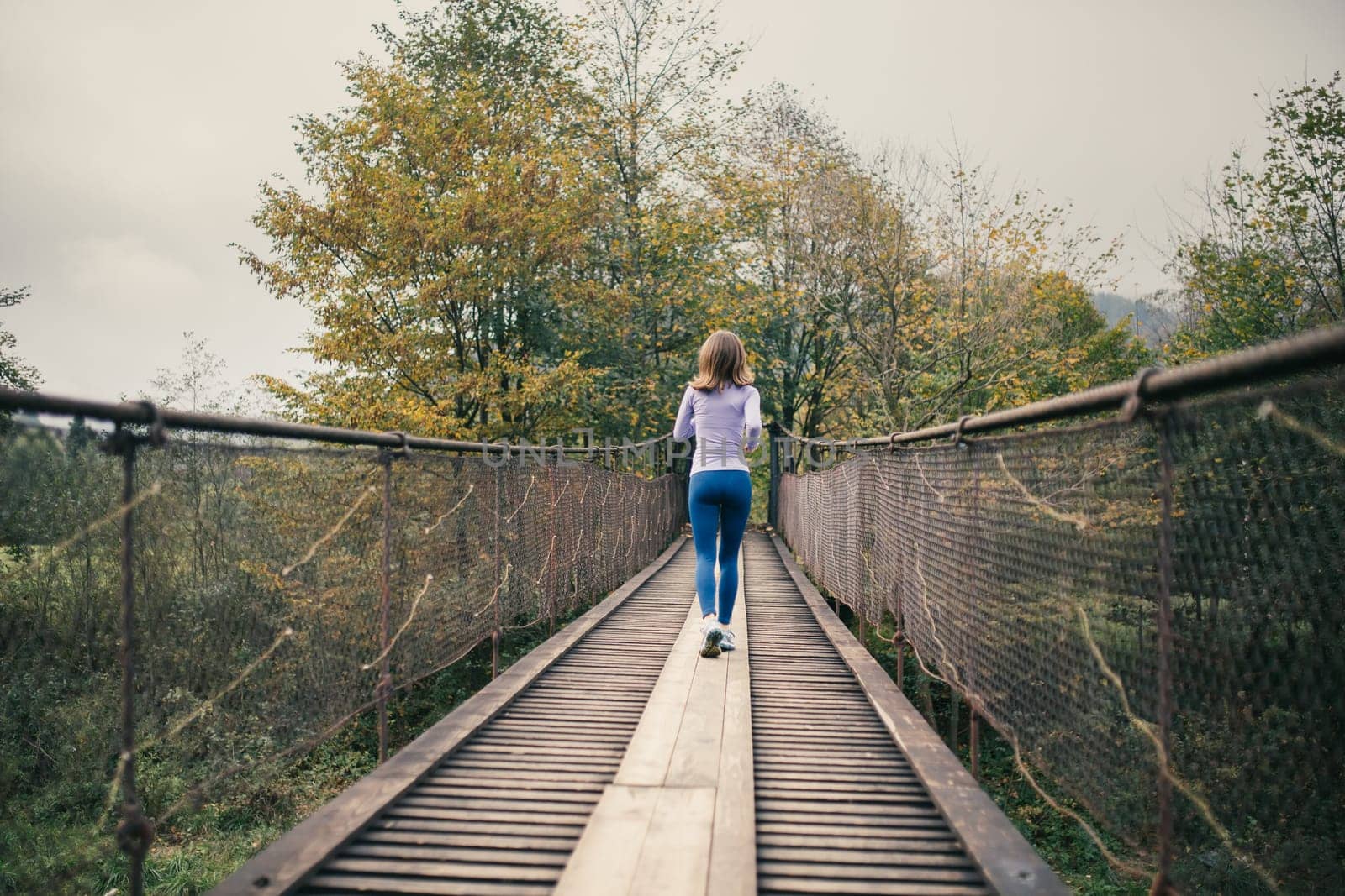 Rear view of a young athletic woman running across a suspension bridge during cardio exercises. Fitness woman working out in nature on a cool autumn day. Concept of active and healthy lifestyle.