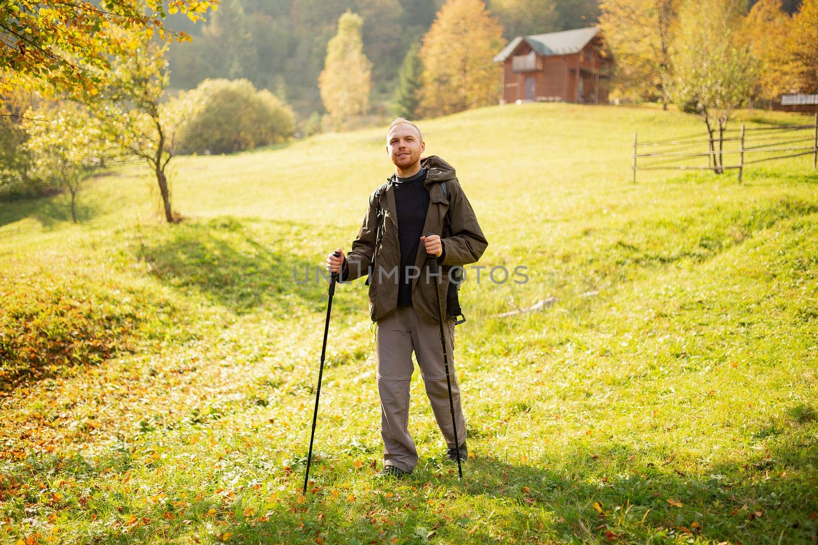 Walking on top of the mountains. Male hiker with hiking equipment stands in the middle of a mountain village on a hill and looks at the camera. Happy man exploring the sunny mountains. Wanderlust.