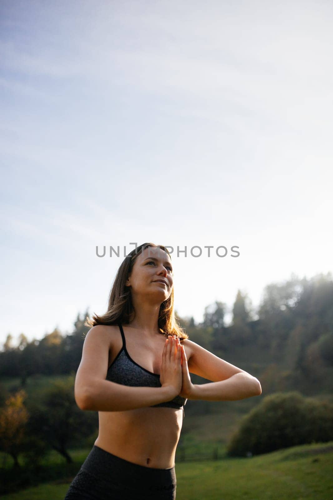 Young woman leading an active lifestyle performs yoga exercises in an open mountain area. Woman puts her palms together and keeps her balance. Daily training concept.