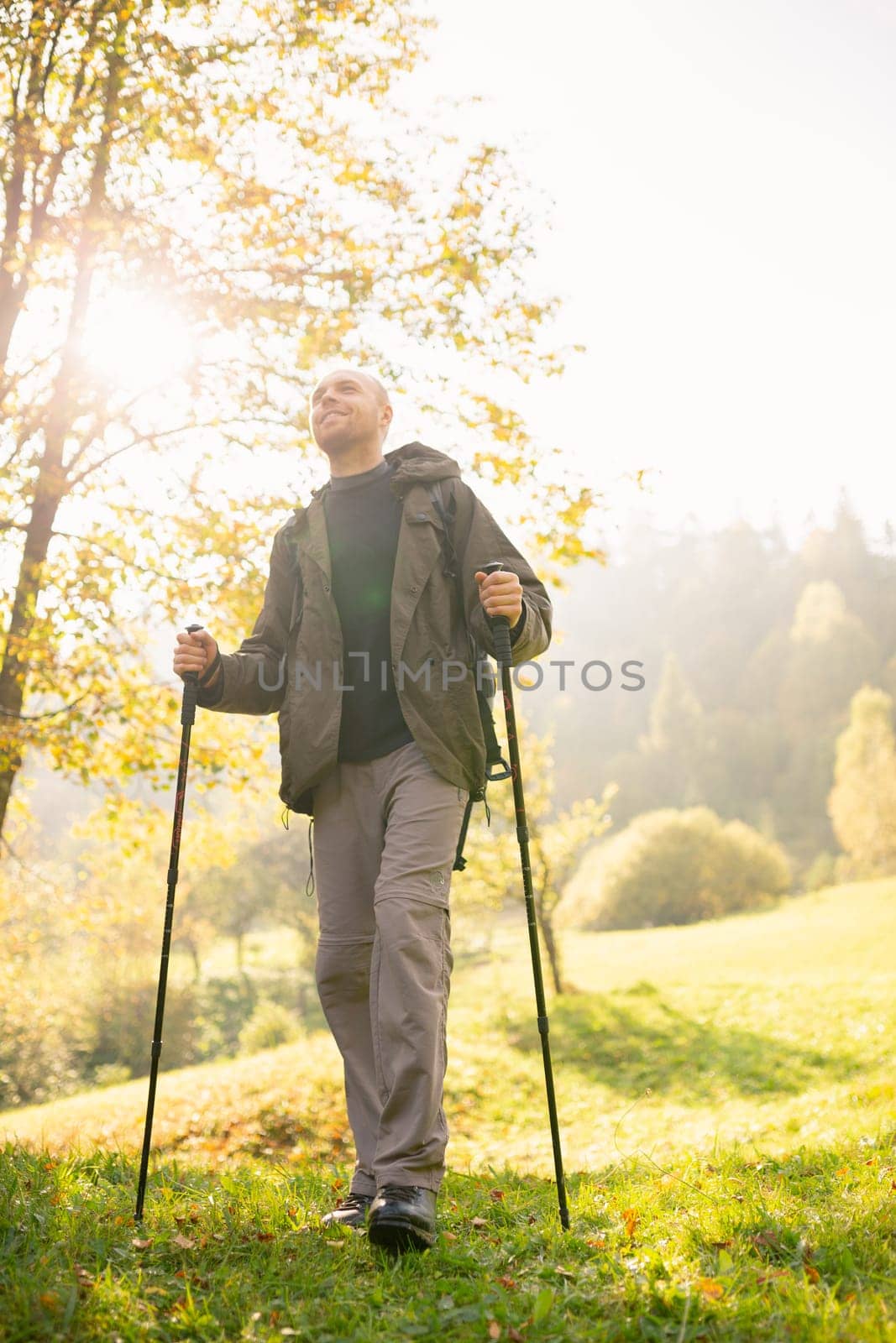 Natural background. View from below of a young male traveler with trekking sticks in his hands standing in the sun. Smiling man enjoys the beauty of nature. Concepts of travel and wanderlust.