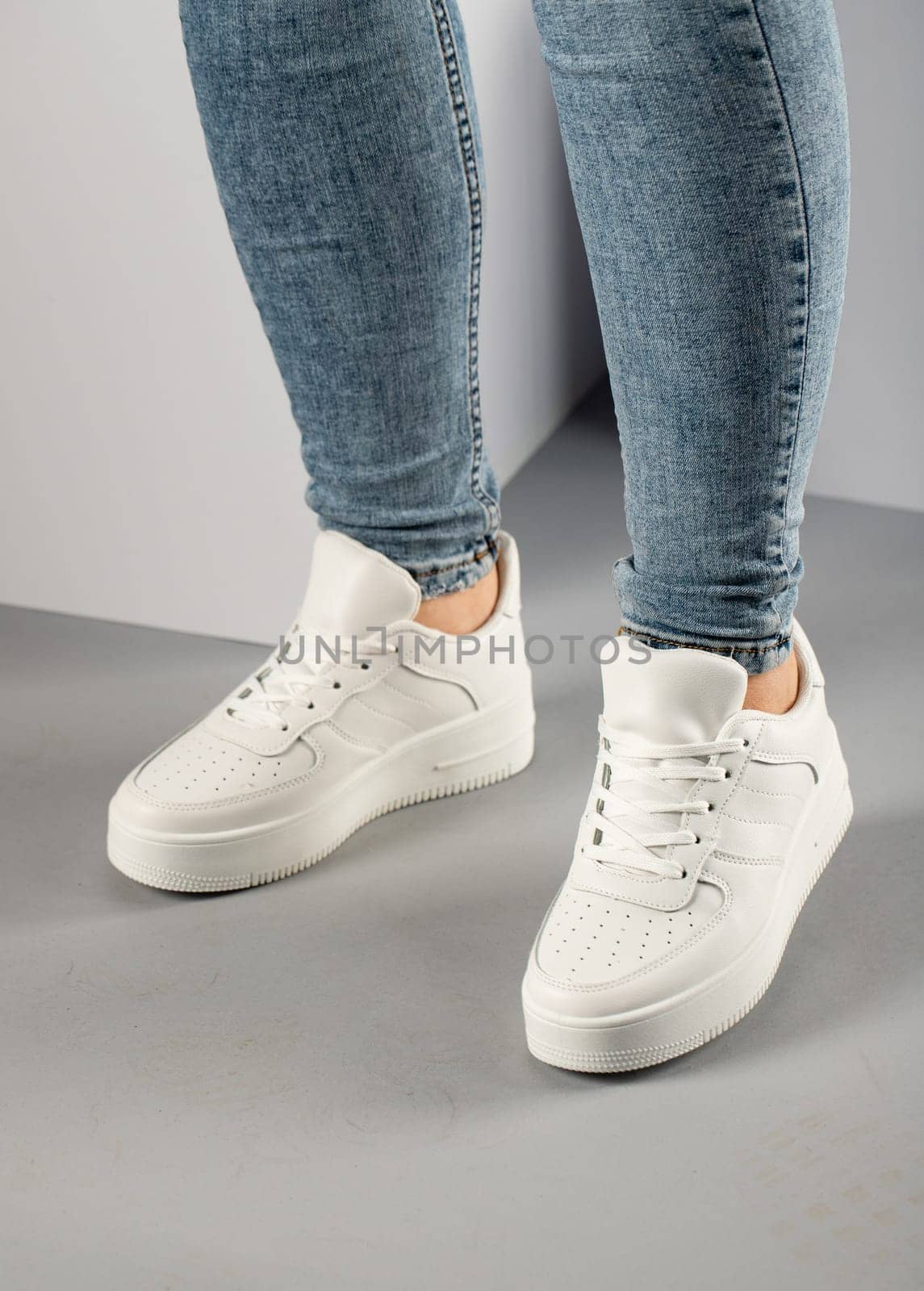 Casual comfortable white sneakers on female model in studio. Sports shoes with laces on feet of unknown young woman who is wearing jeans and standing on gray floor.