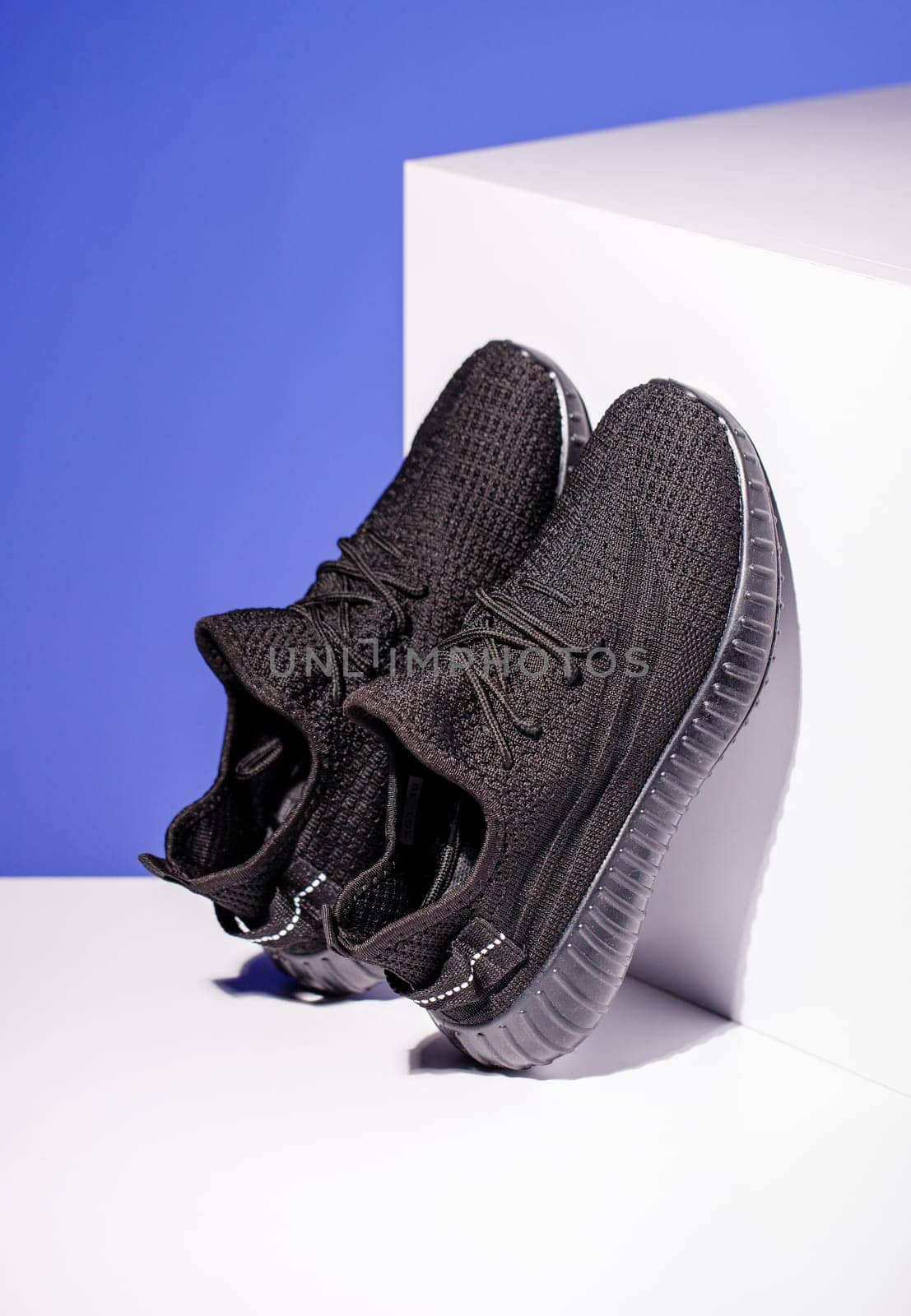 Black sneakers on blue background with white podium in form of geometric cube. Side view of unisex style sneakers standing leaning on podium. Spring and summer fashion concept.