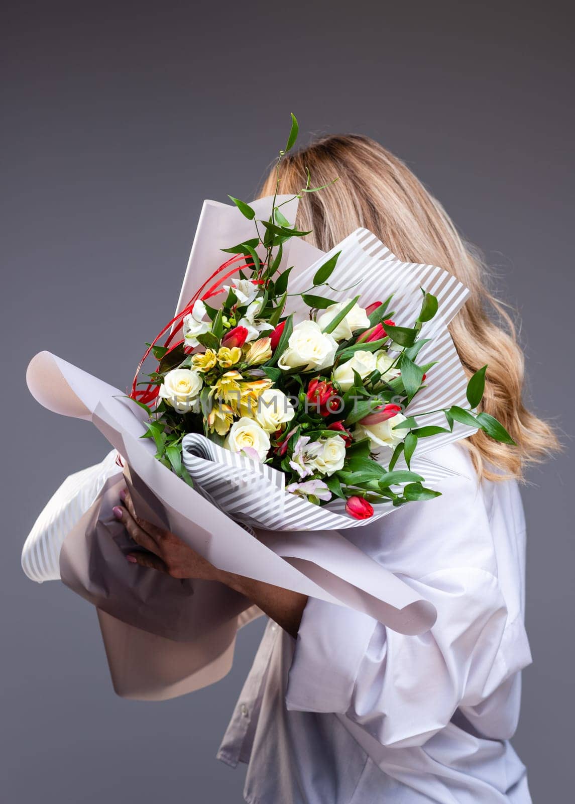 Woman in a delicate white shirt holds a bouquet of flowers with roses, tulips and greenery. Unknown blonde woman stands sideways on a gray background and holds flowers that cover her face.