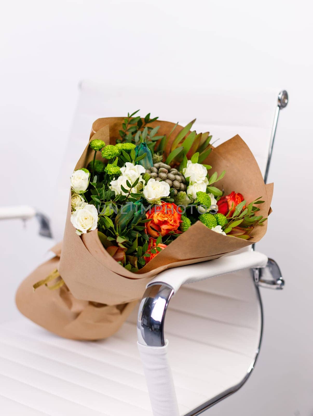 Order and delivery of original bouquet of flowers for any holiday. Bouquet of red, white and green flowers wrapped in brown kraft paper lies on white chair in room. Celebration concept.
