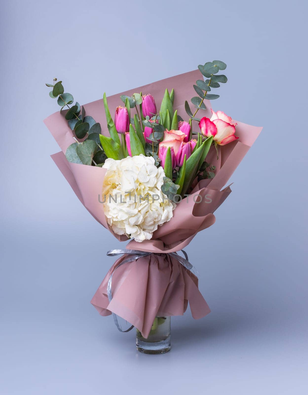Handmade bouquet for Mother's Day in a festive paper package stands in a vase on a blue background. Beautifully packaged flower arrangement with pink tulips, white hydrangea and eucalyptus twigs.