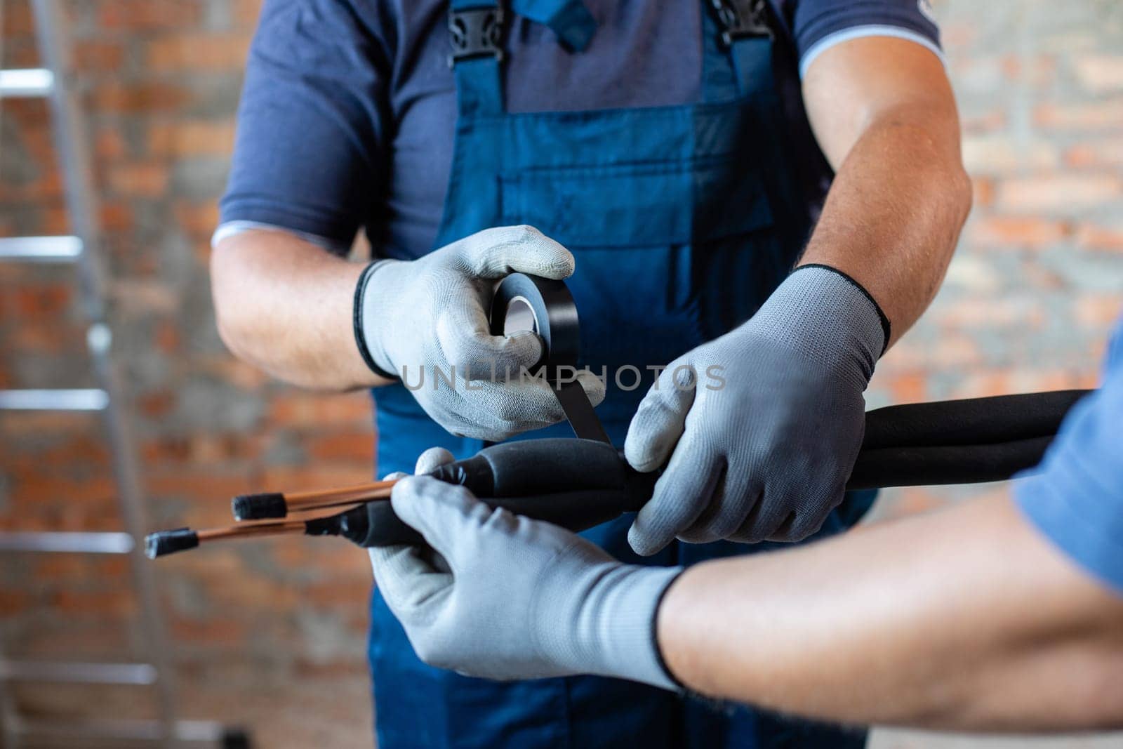 Two adult male workers in overalls are preparing to install underfloor heating pipes. Close up of repairmen hands in work gloves fastening pipes and wires with insulating tape.