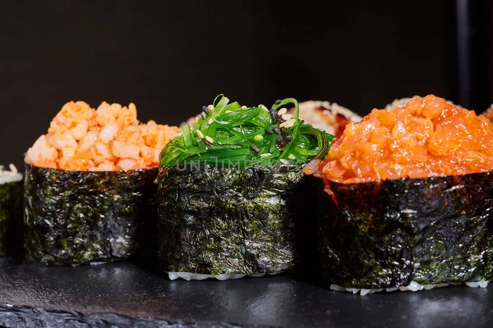 Close up of three pieces of sushi with different fillings on a black plate. Sushi rolls with seaweed, shrimp, salmon and sprinkled with sesame seeds.