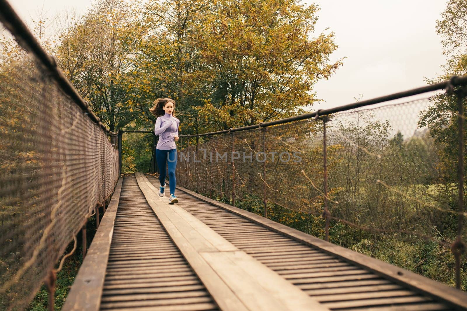 Young woman spends time with benefit. Female runner begins to jog over a wooden suspension bridge in the middle of a mountain forest. Fitness woman working out in nature on a cool autumn day.