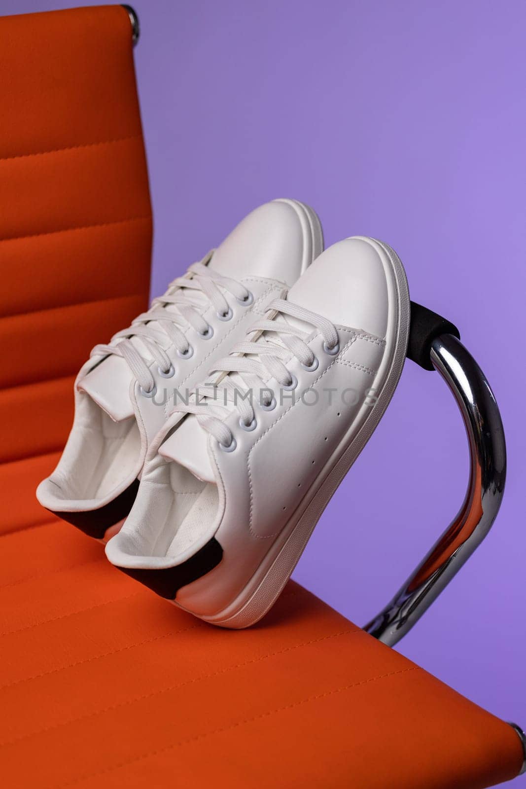 Creative promotion. Advertising photo of white women's or teen sneakers on chair in studio. Close up of white sneakers with black inserts standing leaning on handle of red chair on purple background.