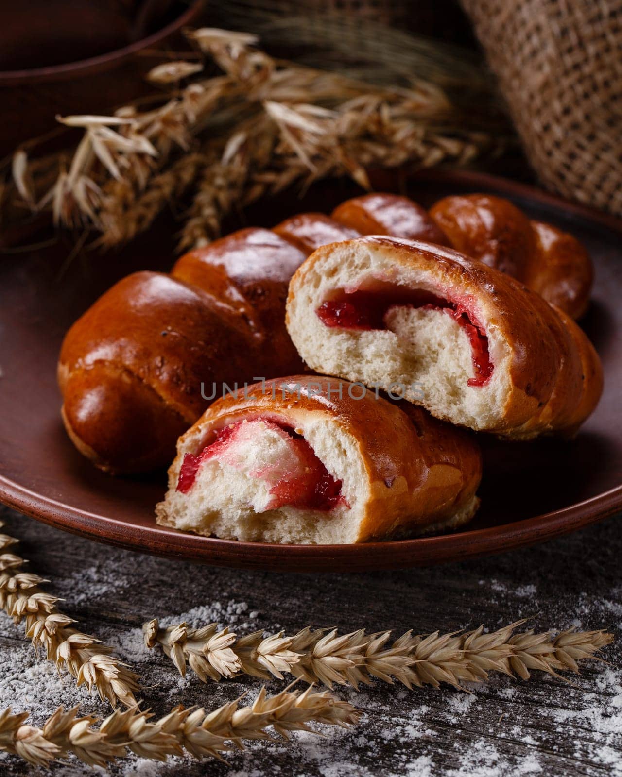 Close up of sweet pastries lying on clay plate on rustic table. Broken bun with cherry or strawberry filling lies next to burlap and ears of wheat on dark wooden table sprinkled with flour.