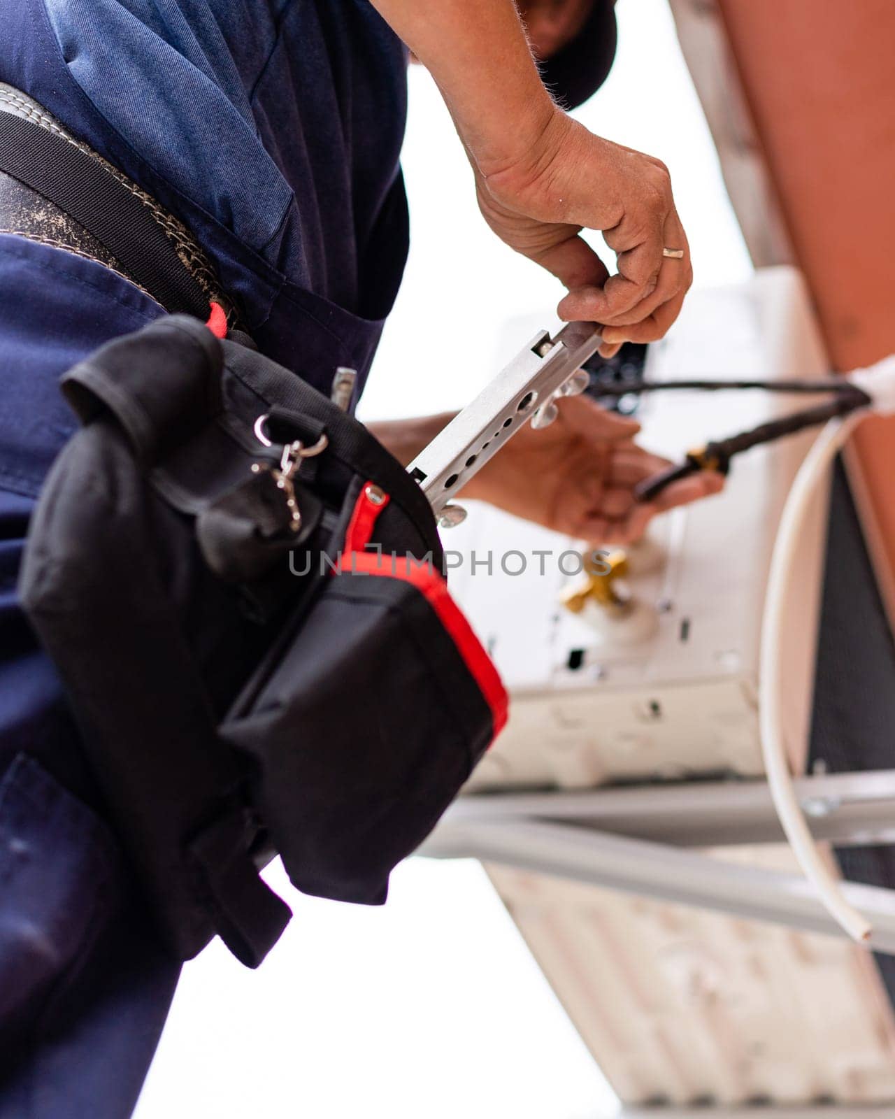 Male technician connects outdoor air conditioning unit to wall of old apartment building. Close-up of worker pulling tool out of his waist bag. Selective fiocus.