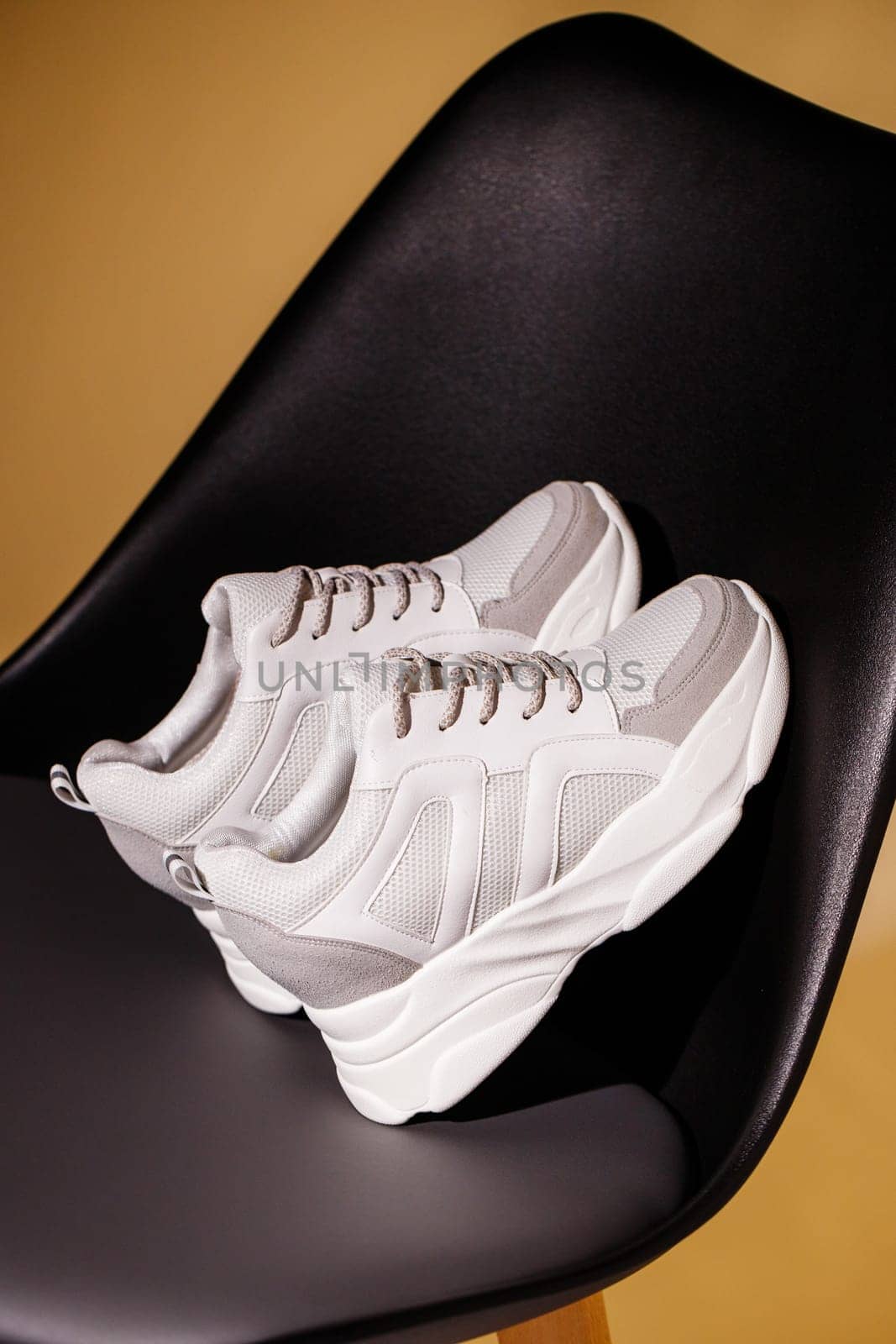 Promotion. Close up of white sneakers leaning on back of black chair on beige studio background. Sneakers are very popular among teenagers or women because of their simple and fashionable appearance.