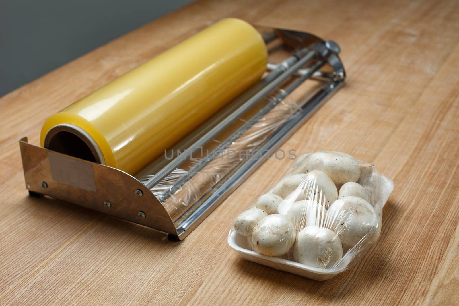 Close up of champignons on plastic tray packed in transparent cling film. Fresh packaged mushrooms on table near large roll of food packaging film. Concept of storing fresh food in supermarkets.