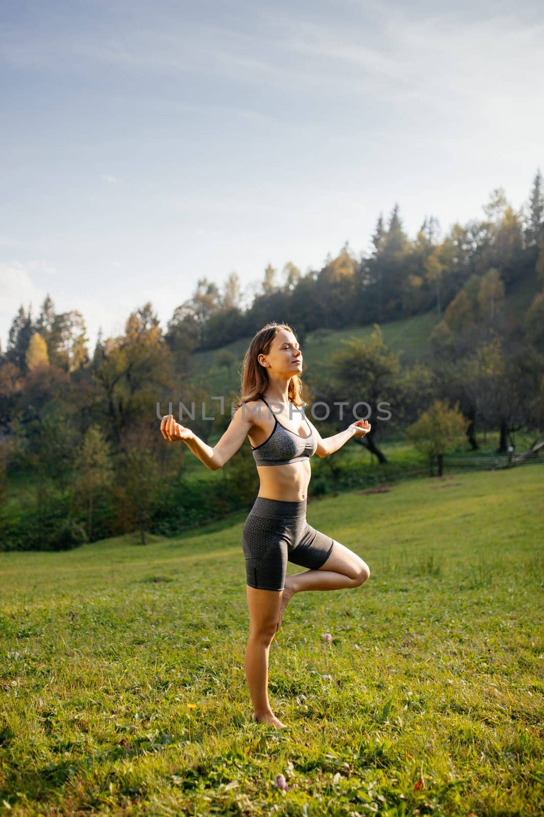 Yoga outdoor. Cute slender young woman with closed eyes meditates, does breathing exercises and keeps balance on one leg standing barefoot on the grass outdoors. Concept of physical and mental health.