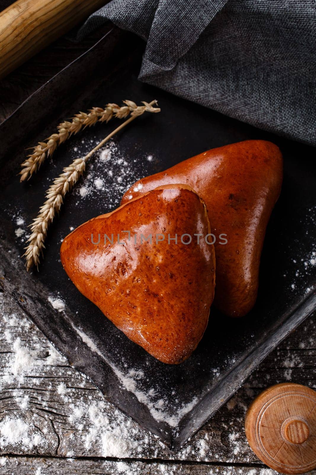 Close up of two fresh baked buns on dark baking plate. Top view of buns lying near burlap and ears of wheat on wooden table sprinkled with flour. Baking concept with rye and flour.