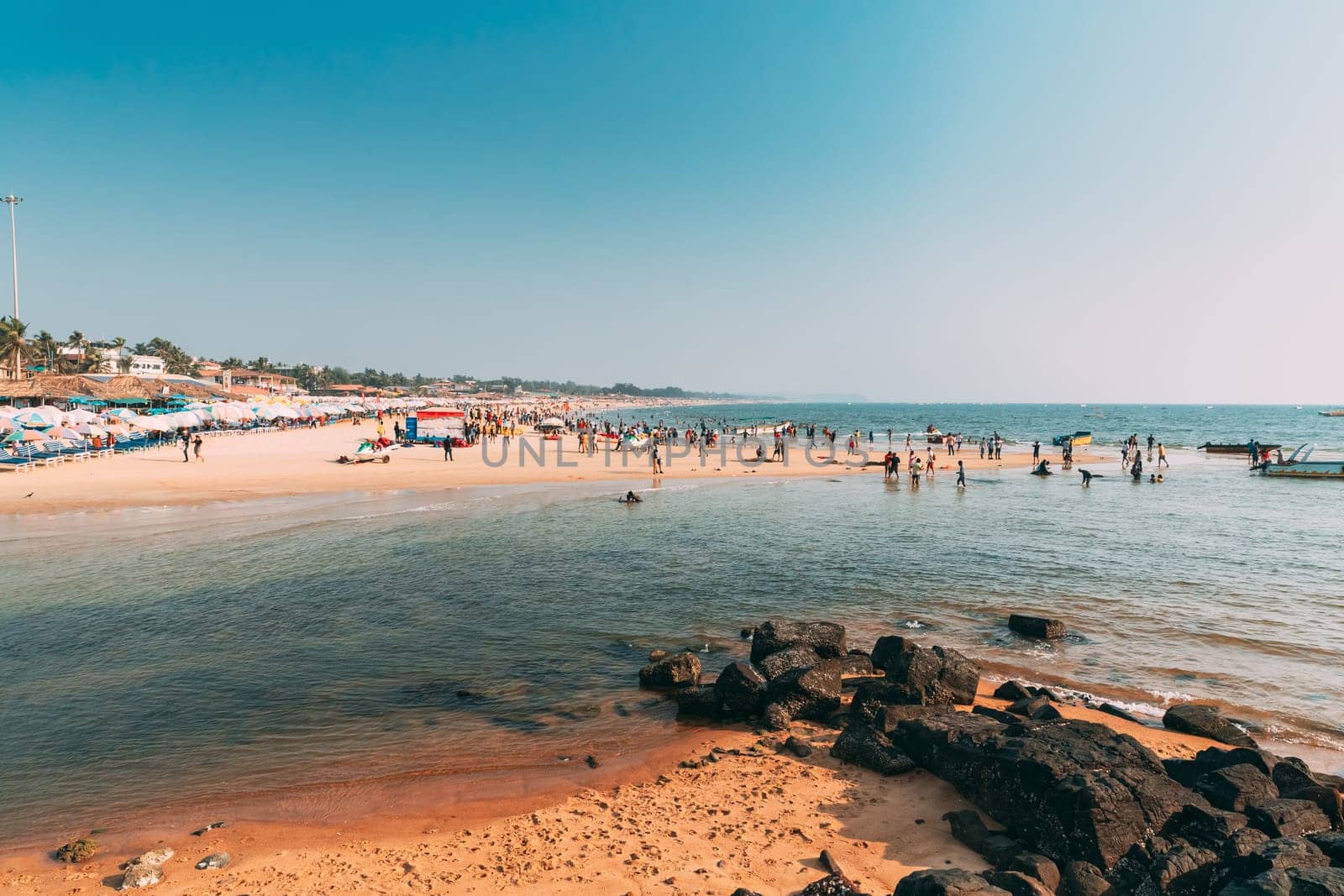 Mapusa, Anjuna, Goa, India. People Visit And Relax On Baga Beach At Sunny Evening Under Blue Sky