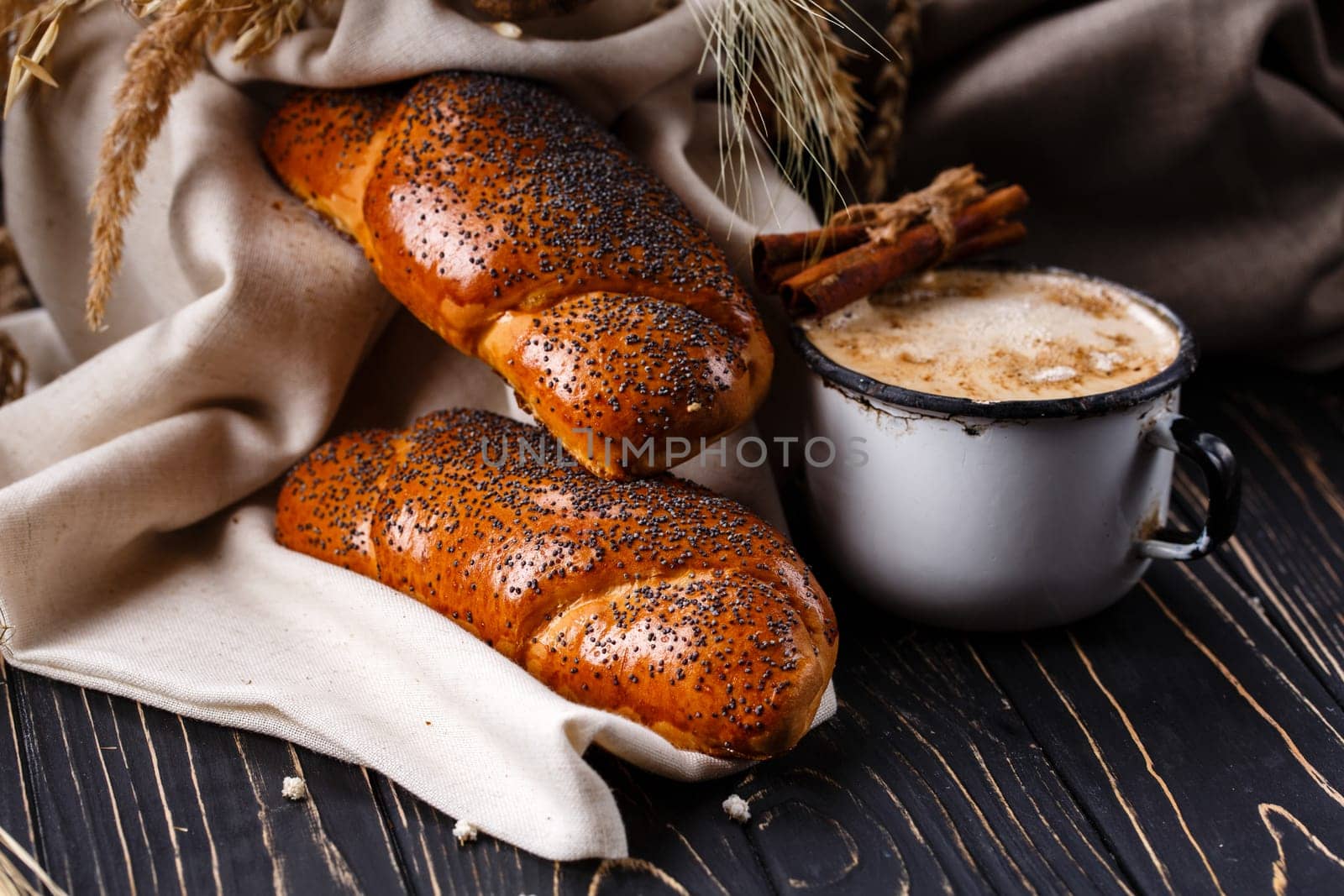Morning energy. Traditional breakfast with poppy seed rolls and coffee on rustic dark wooden table. Close up of two fresh ruddy buns on burlap near old enamel mug with coffee and cinnamon sticks.