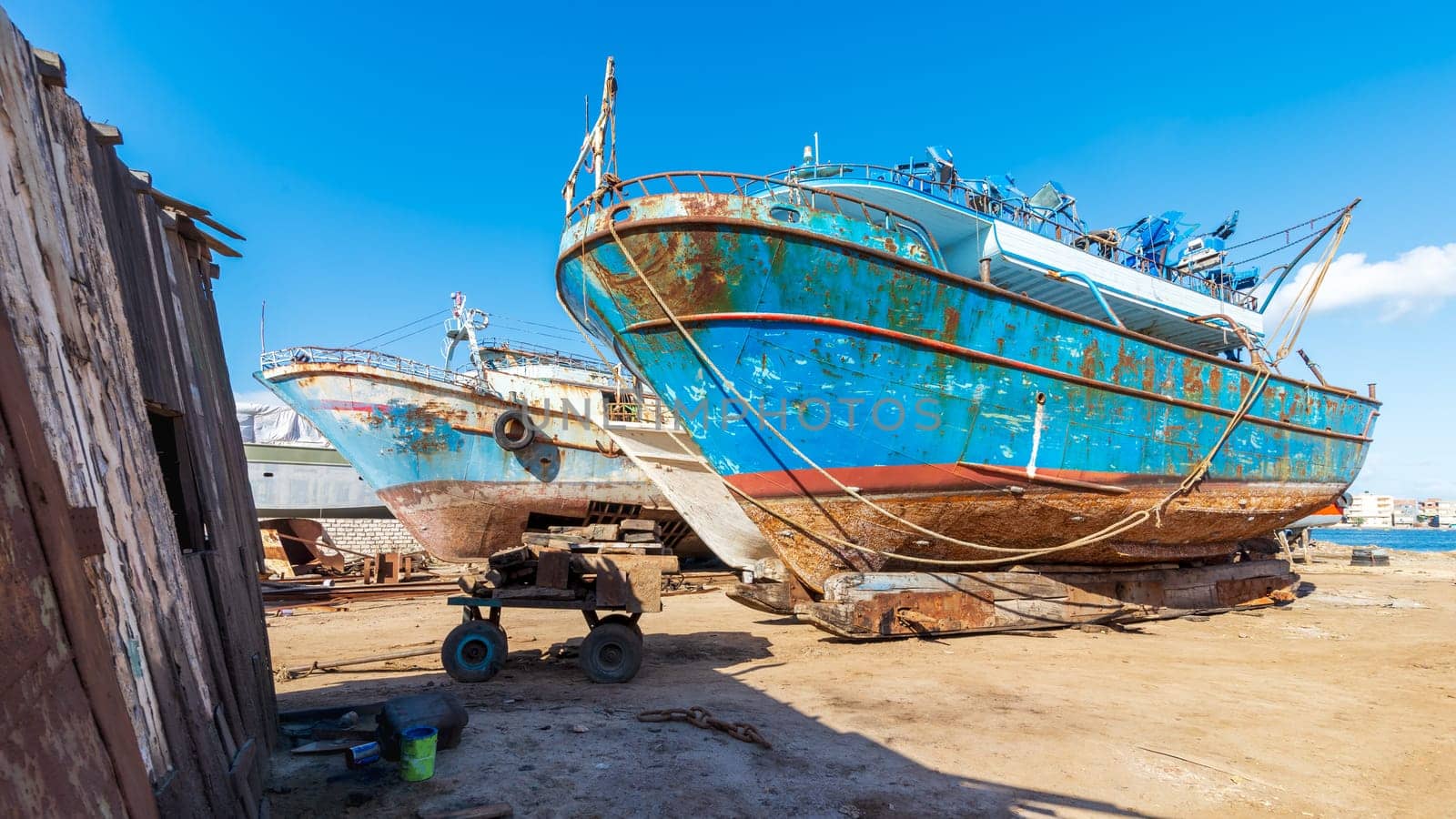 Old rusty vessels under repairing located on grungy dry dock in shipyard against blue sky on sunny day in old shipbuilding plant