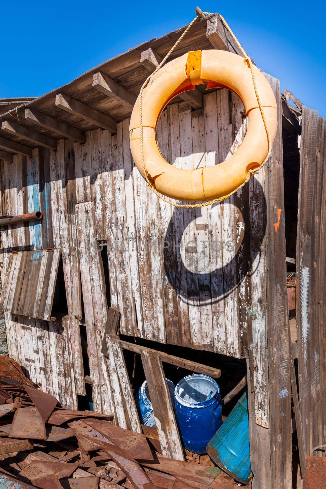Old orange life saver hanged on edge of roof of shabby small wooden building among lot of boards and clear blue sky