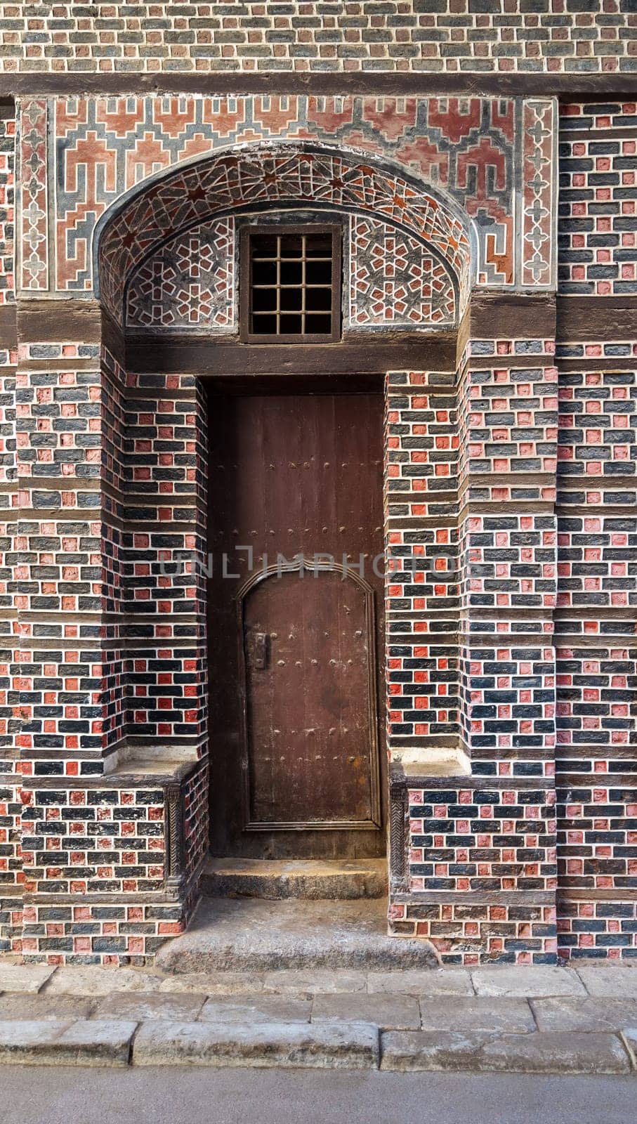 Wooden grunge decorated arched entrance gate with inner small wooden door on wall with black and red bricks with white seam