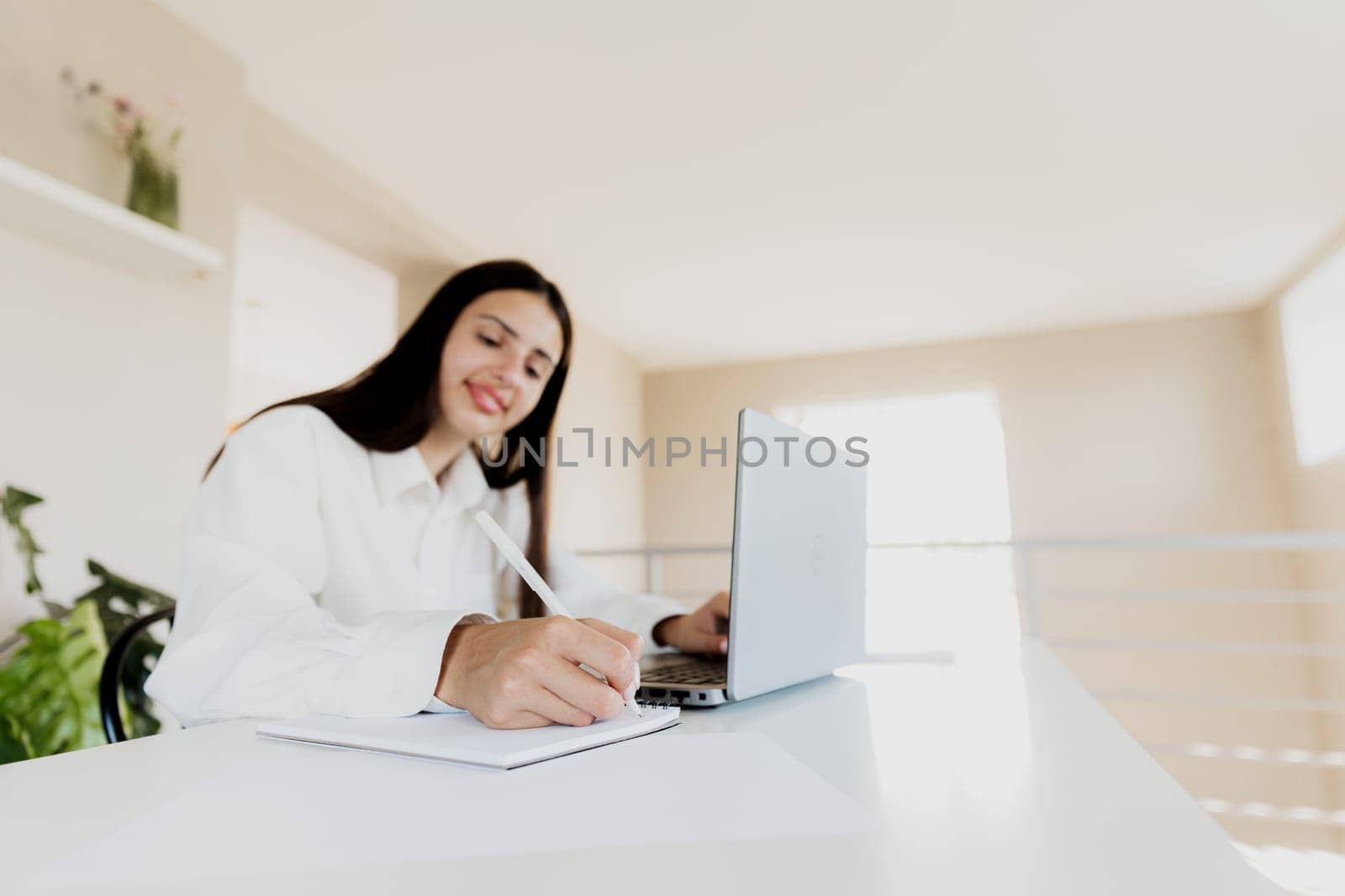 Caucasian young business woman sitting at desk, reading from computer, taking notes at her white notebook. Student working from home during online work meeting or webinar. Happy smiling lady at office