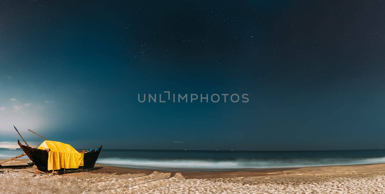 Goa, India. Real Night Sky Stars. Natural Starry Sky Blue Color Above Sea Seascape Ocean Beach. Background. Parked Old Wooden Boat At Coast. Panorama, Panoramic View.