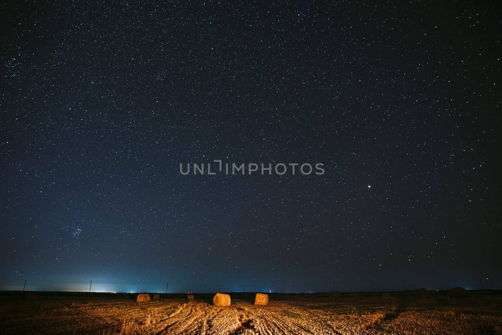 Night Starry Sky Above Haystacks In Summer Agricultural Field. Night Stars Above Rural Landscape With Hay Bales After Harvest. Agricultural Concept.