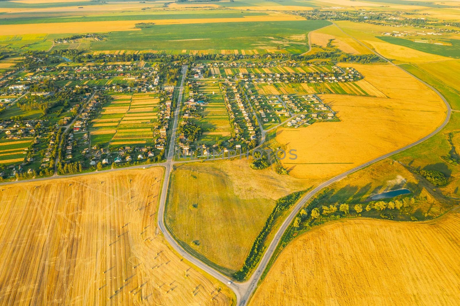 Aerial View Of Village And Hay Rolls, Straw Field Landscape In Summer Evening. Haystack, Hay Roll in Sunrise Time. Agricultural Countryside landscape In Harvest Season