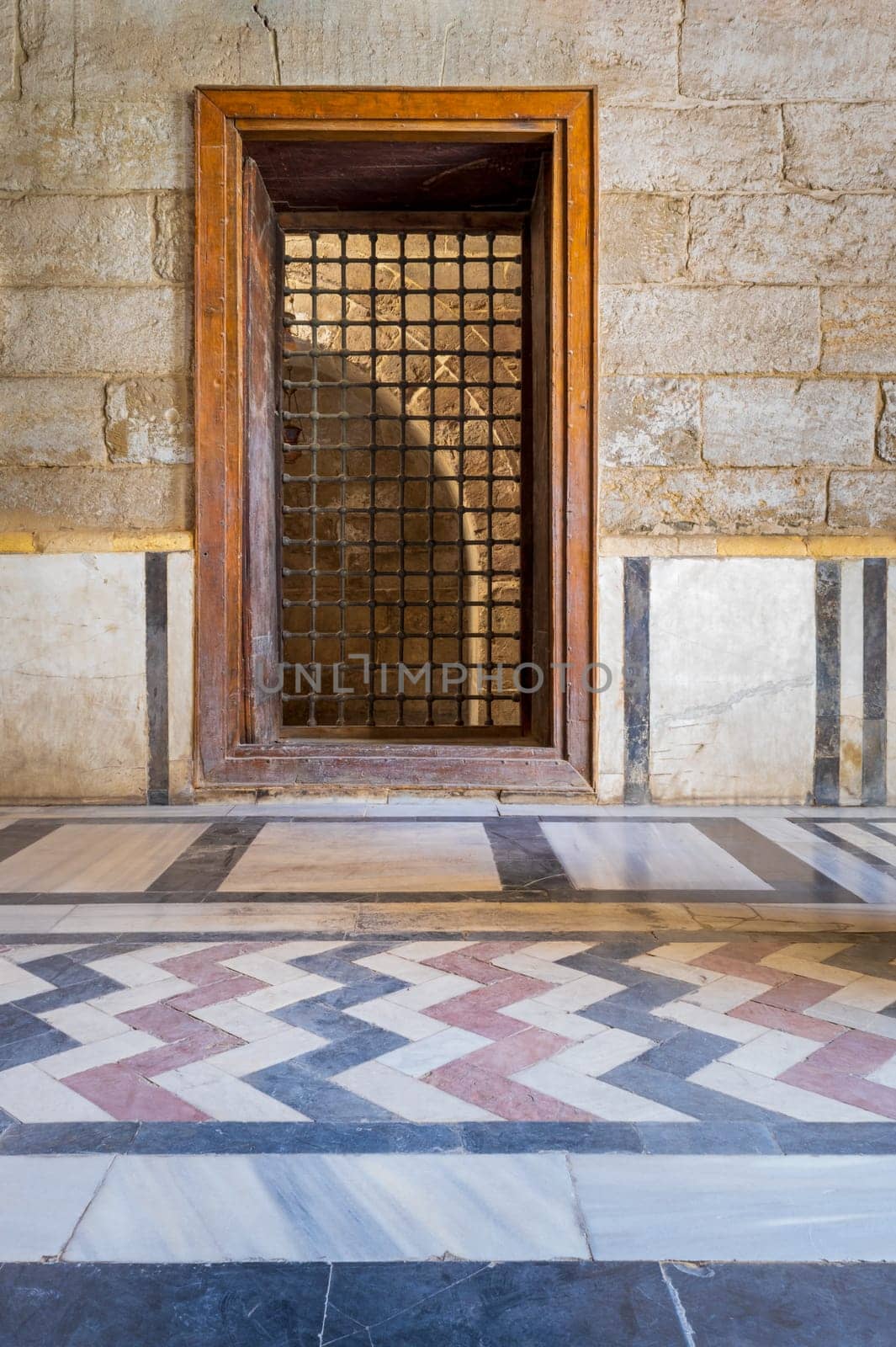 Recessed wooden window with decorated iron grid over stone bricks wall and decorative colorful floor at public historic mosque of Sultan Barquq, El Moez Street, Old Cairo, Egypt