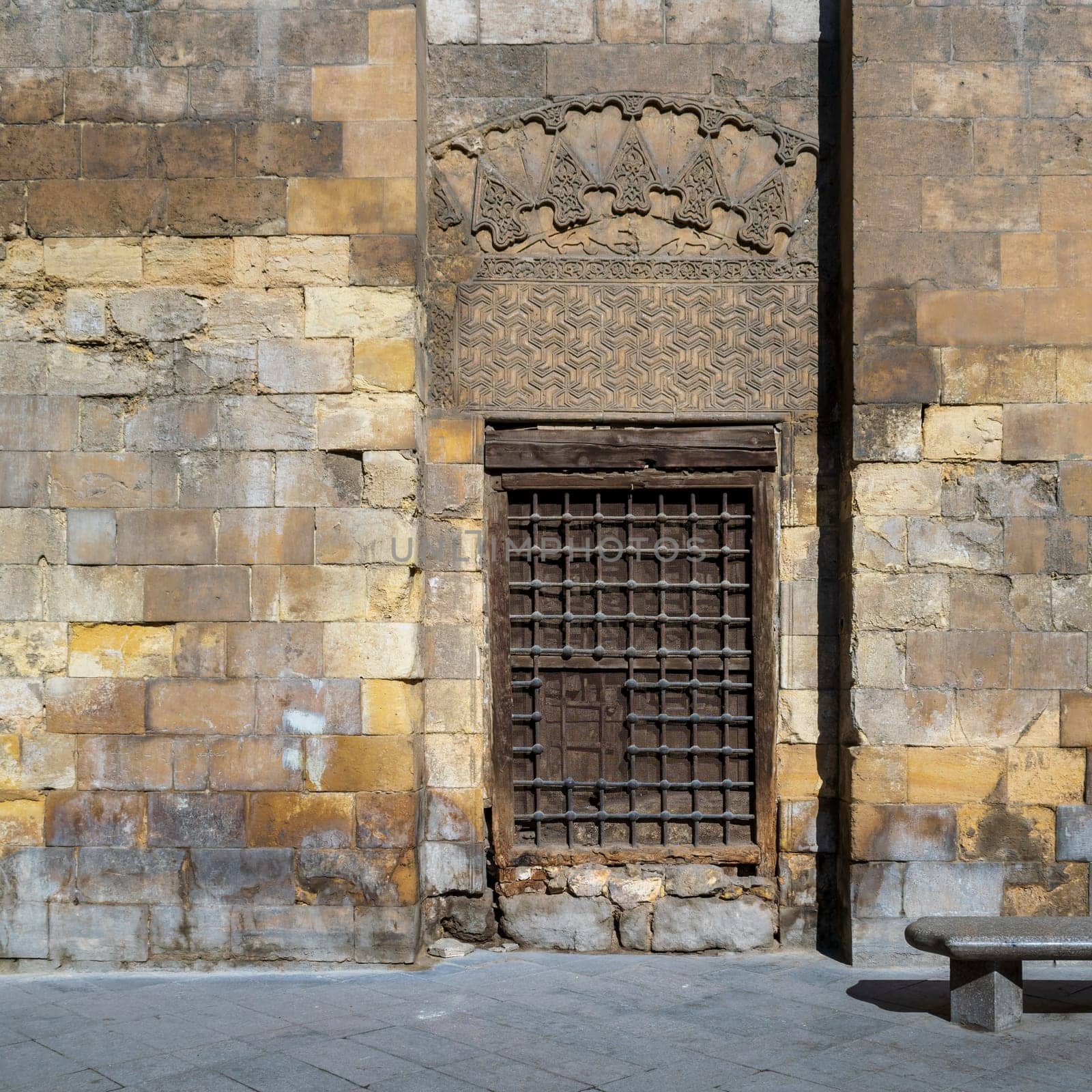 Daylight shot of grunge wooden window with decorated iron grid over stone bricks wall and marble garden bench, Moez Street, Cairo, Egypt