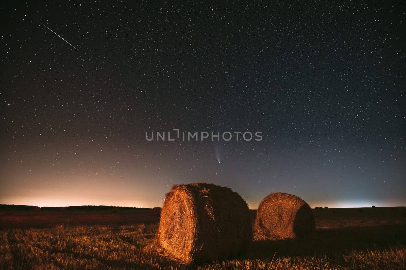 Meteor And Comet Neowise C2020 F3 In Night Starry Sky Above Haystacks In Summer Agricultural Field. Night Stars Above Rural Landscape With Hay Bales After Harvest.