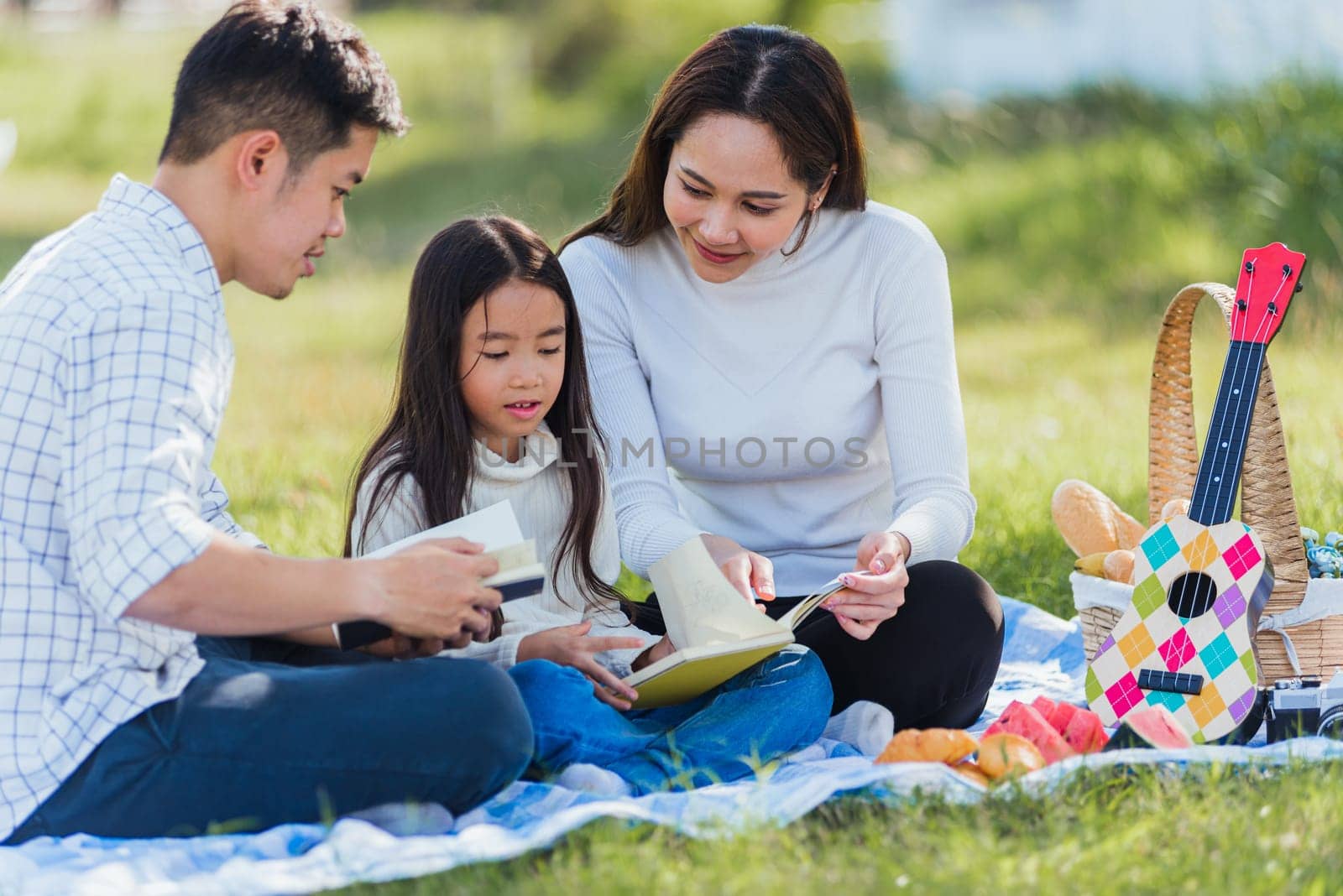 Happy Asian young family father, mother and child little girl having fun and enjoying outdoor on picnic blanket reading book in park at sunny time, summer leisure spring concept